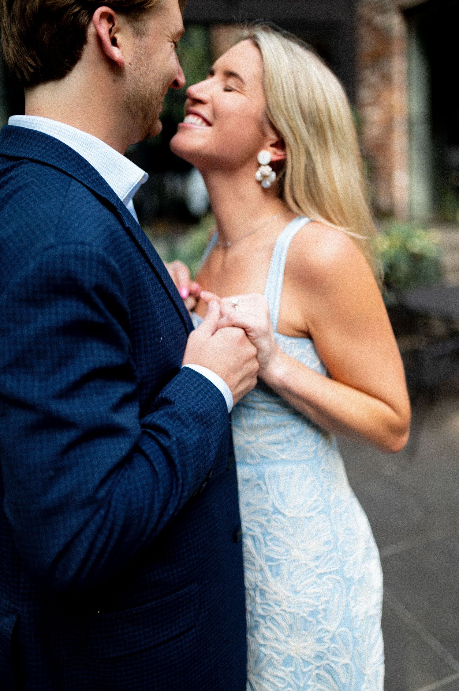 A smiling blonde woman in a light blue dress holds hands with a man in a navy suit during a romantic outdoor moment.