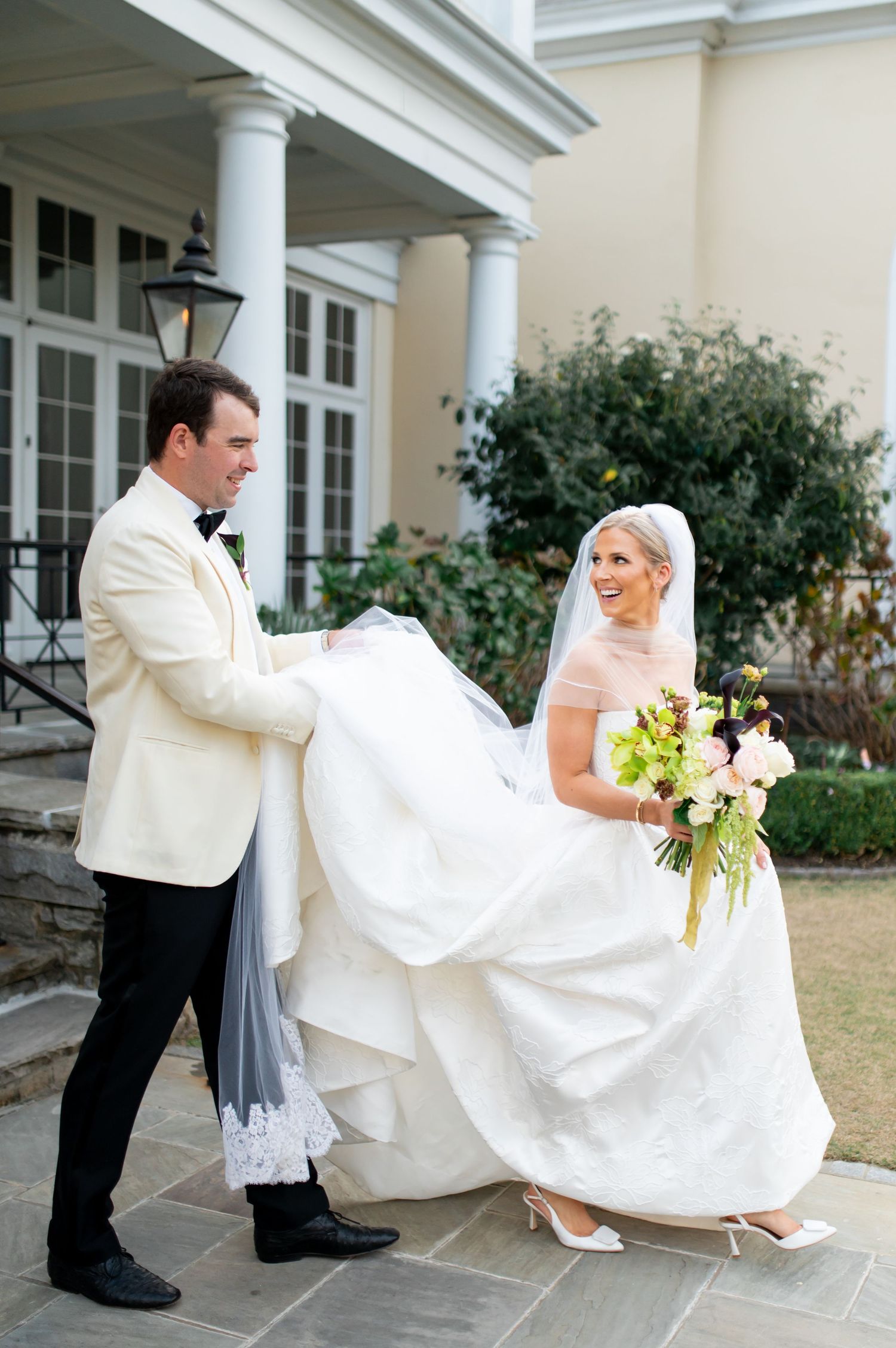 Elegant wedding couple posing outdoors near white columns with flowing dress movement.