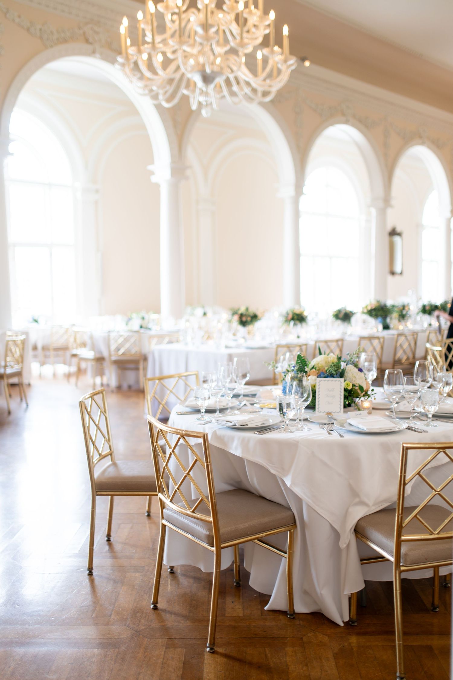 Elegant ballroom with white tables, gold chairs, and crystal chandeliers set for a wedding reception.