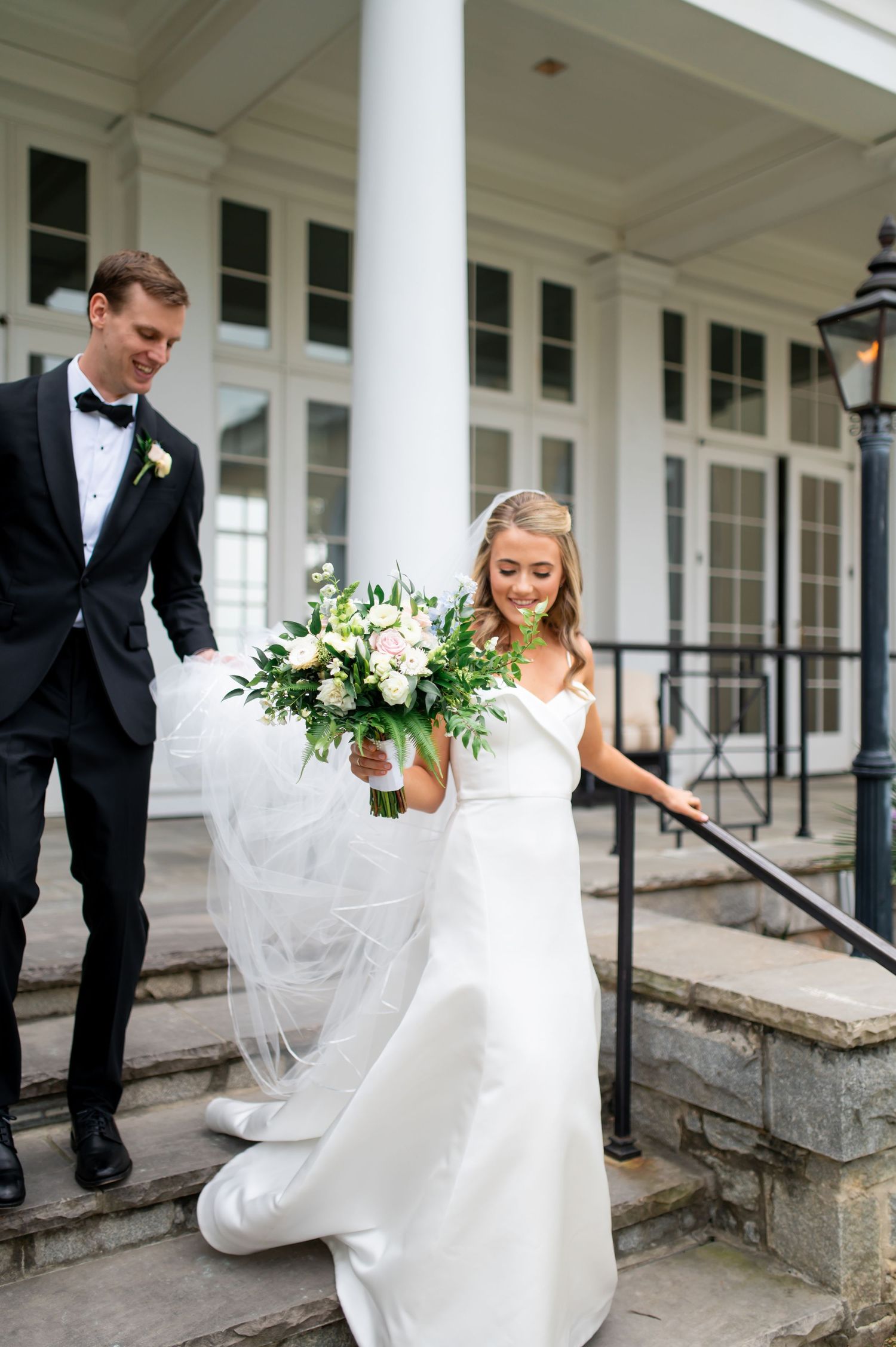 Wedding couple walking along a white columned porch with flowing dress and floral bouquet.