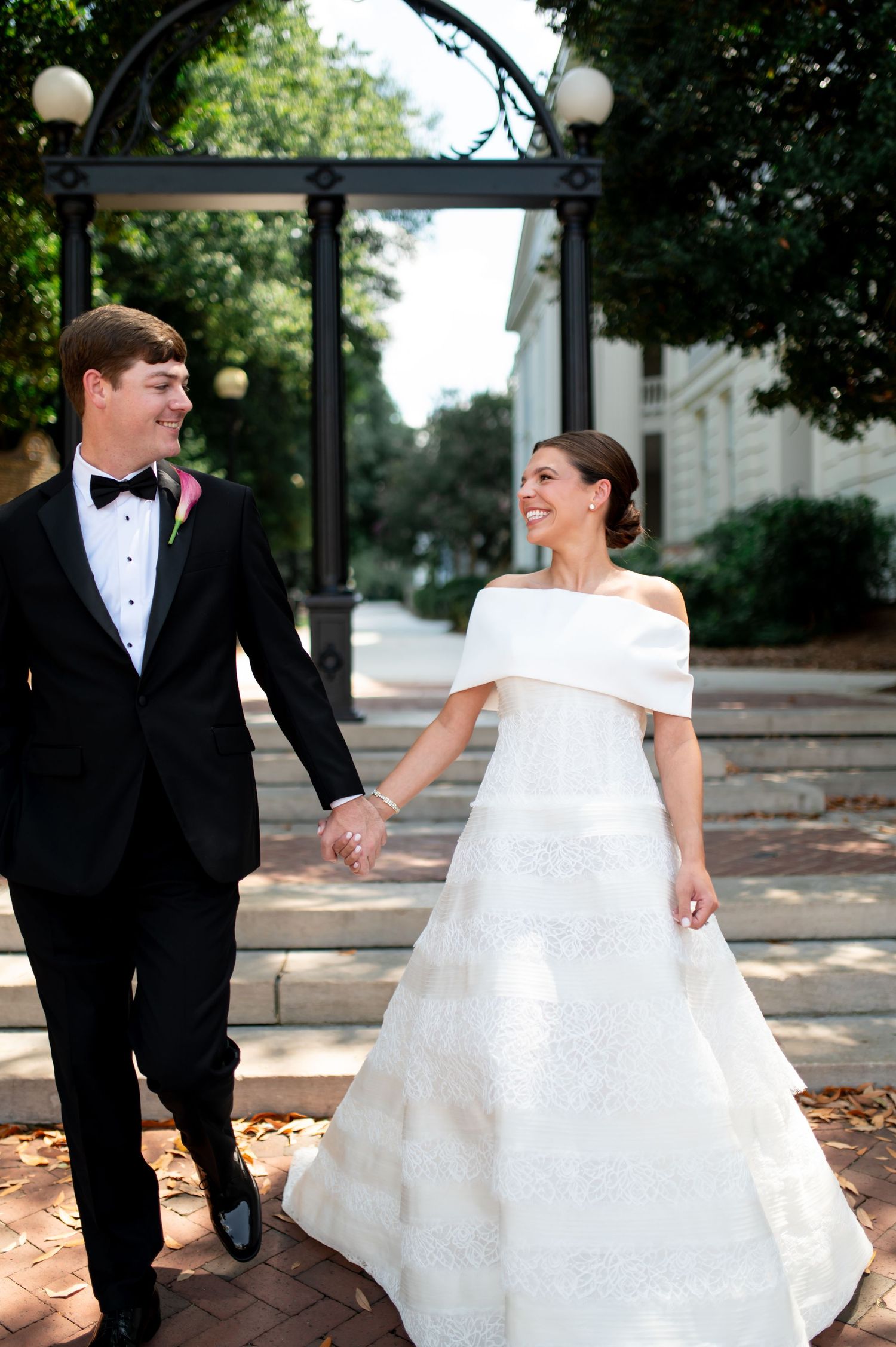 Bride and groom hold hands walking past ornate iron gate, she wears off-shoulder lace gown, he wears classic black tuxedo.