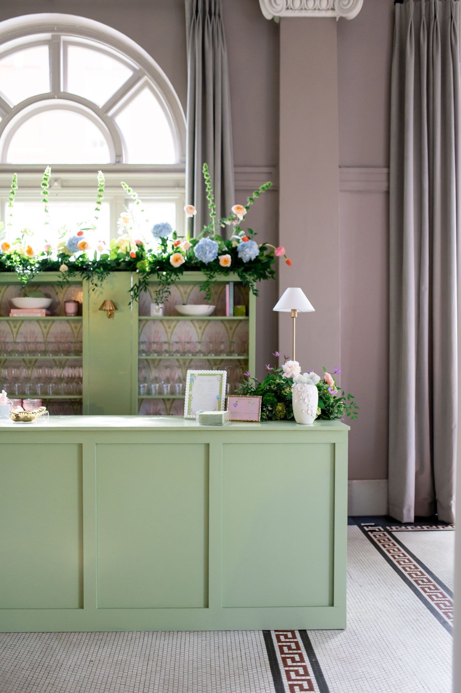 Elegant green reception desk adorned with colorful floral garland near arched window in venue.