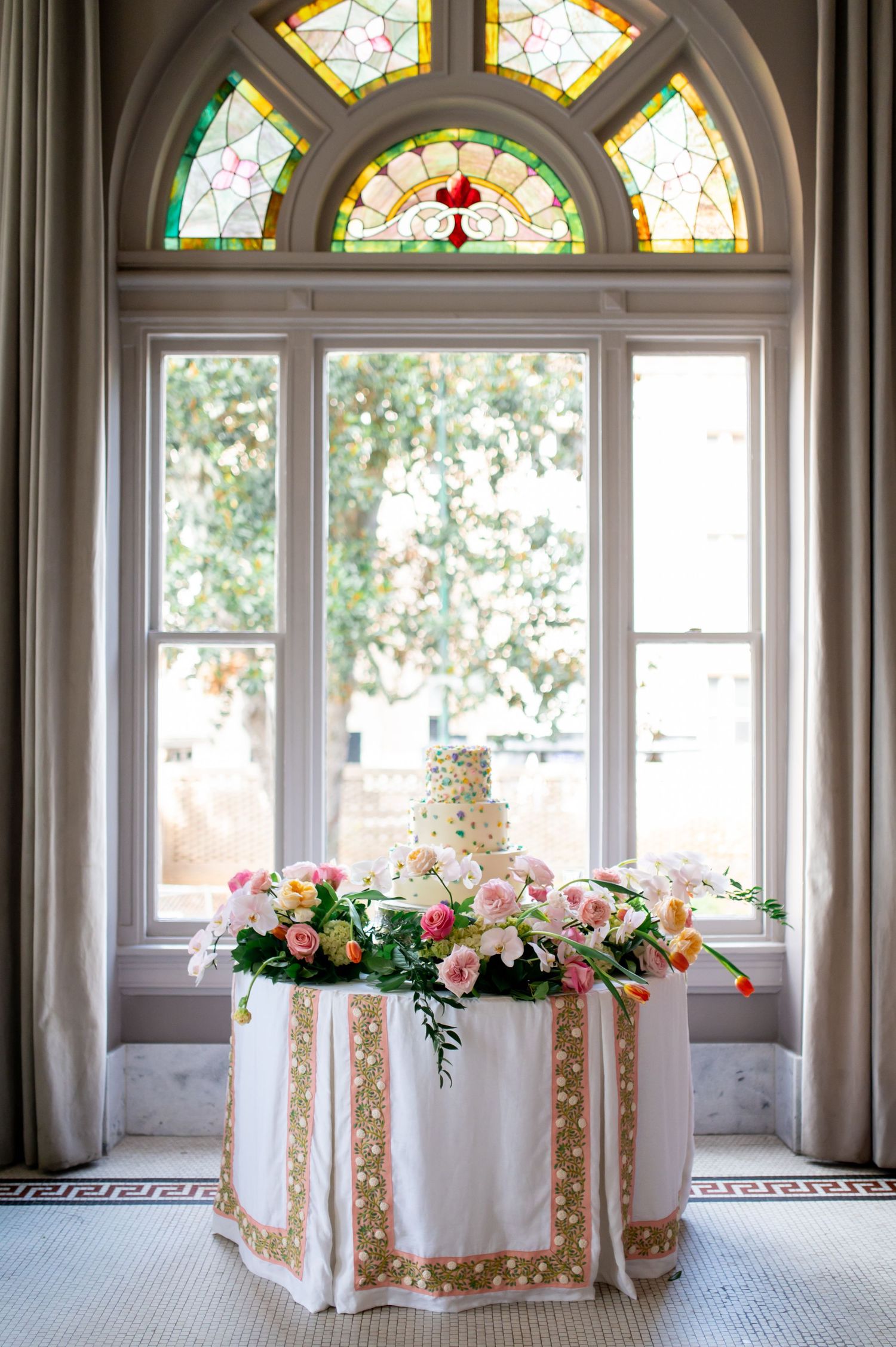 Multi-tier wedding cake on floral-decorated table beneath stunning stained glass arched window.