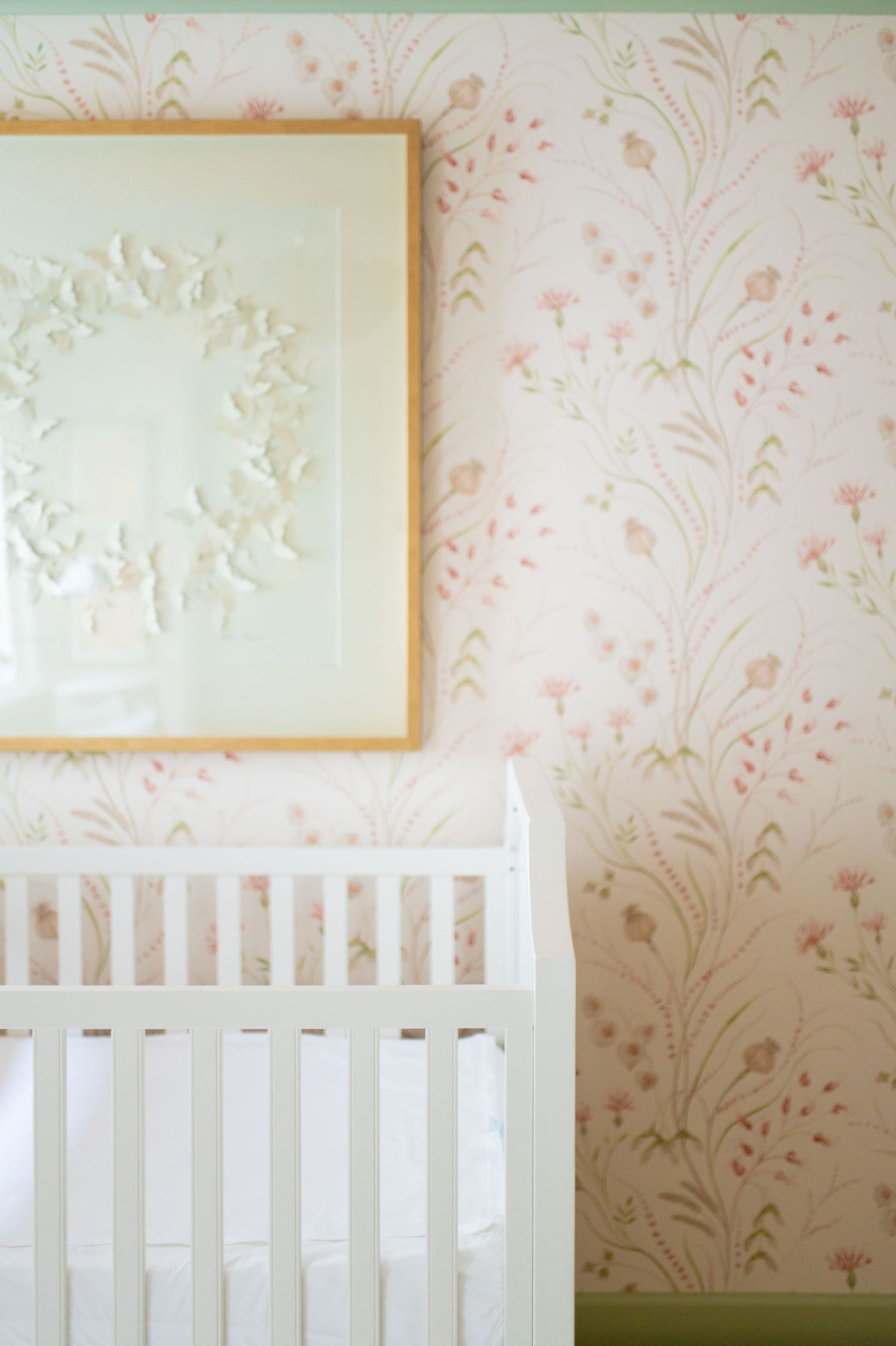 White crib against pink floral wallpaper in a nursery with decorative artwork mounted above.