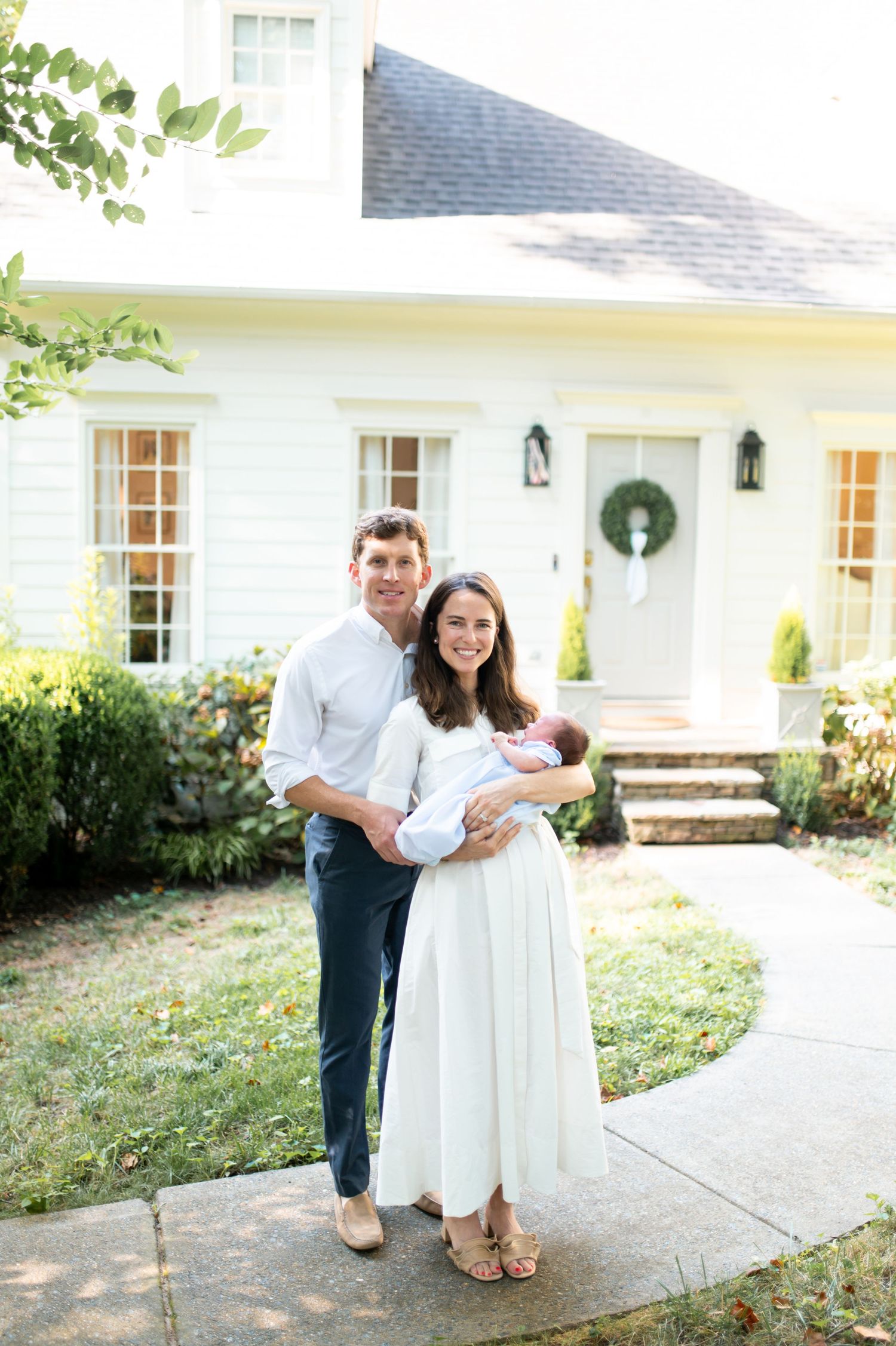 A couple poses lovingly together outside a white house with black shutters and landscaped garden.
