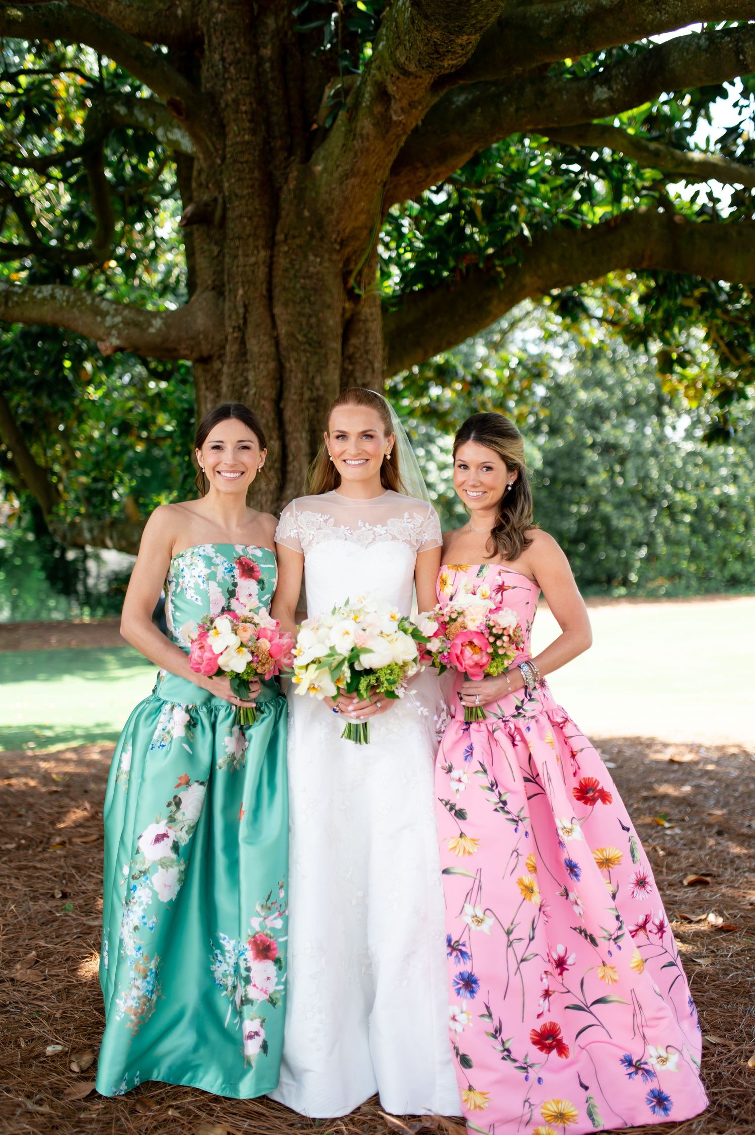 Bride poses with two bridesmaids in colorful floral strapless gowns under large oak tree.