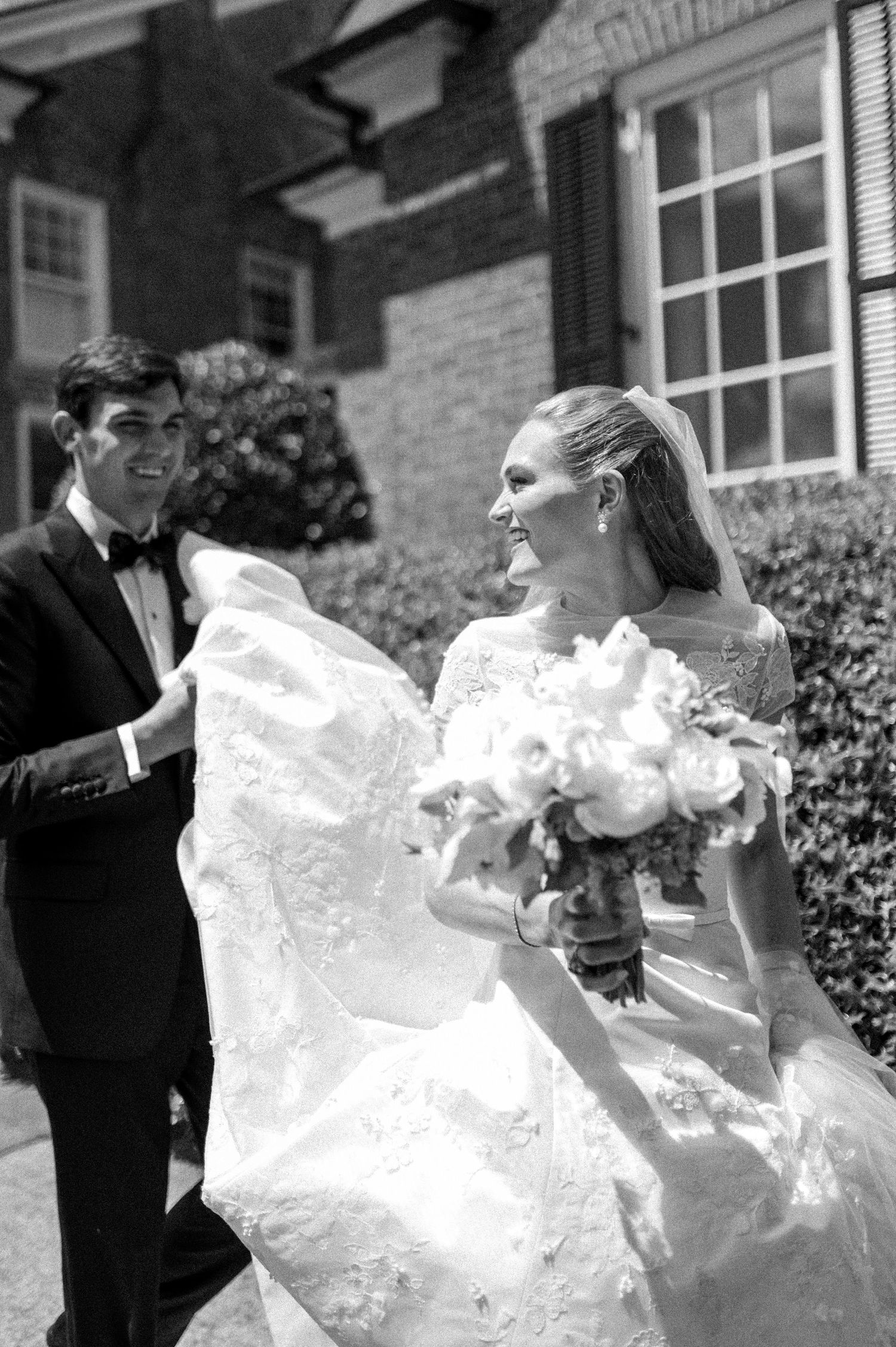 Black and white photo of groom helping bride with flowing lace wedding gown outside brick building.