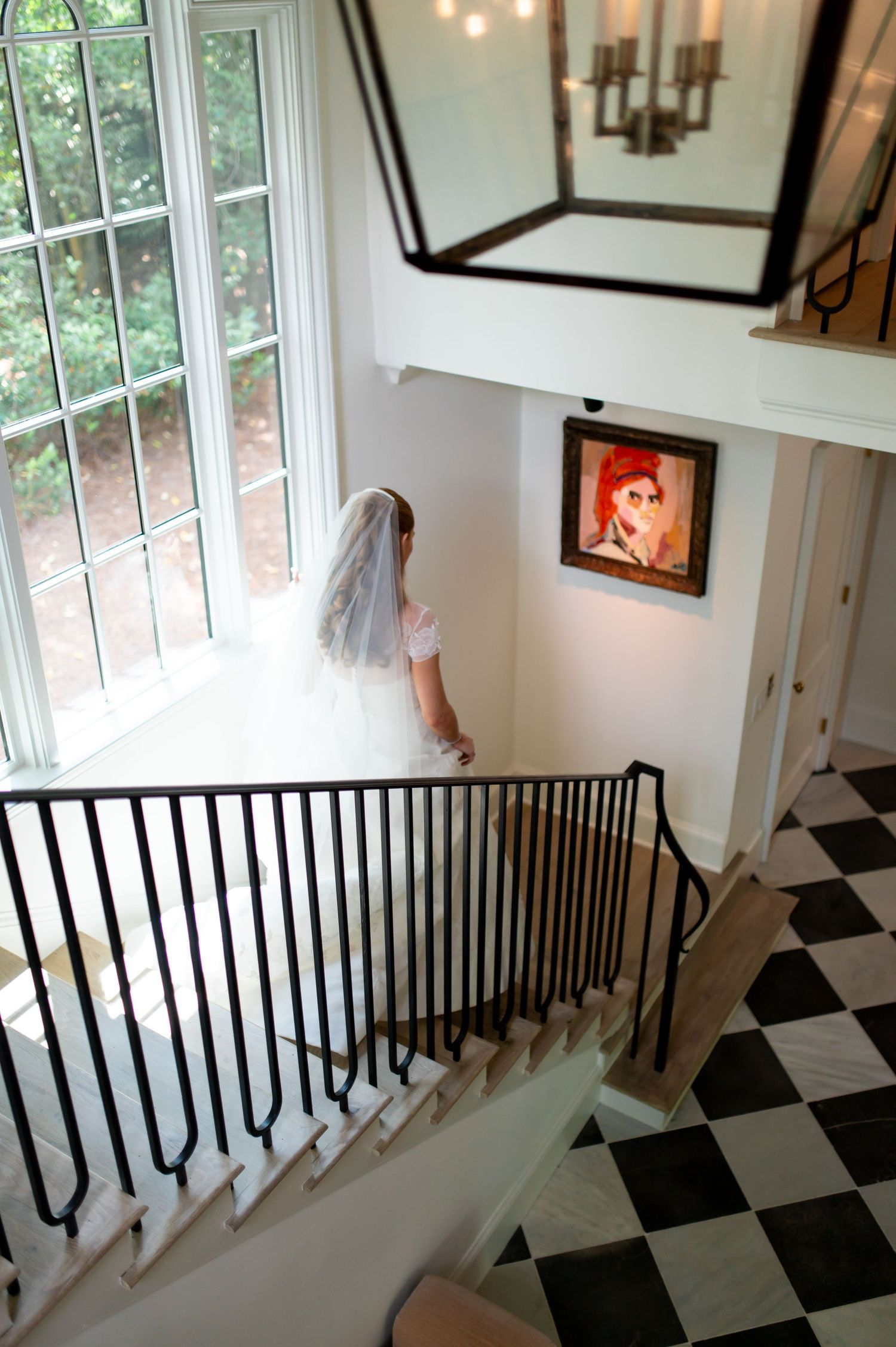 Bride in white gown and veil descends elegant staircase with black iron railing and checkered marble floor.