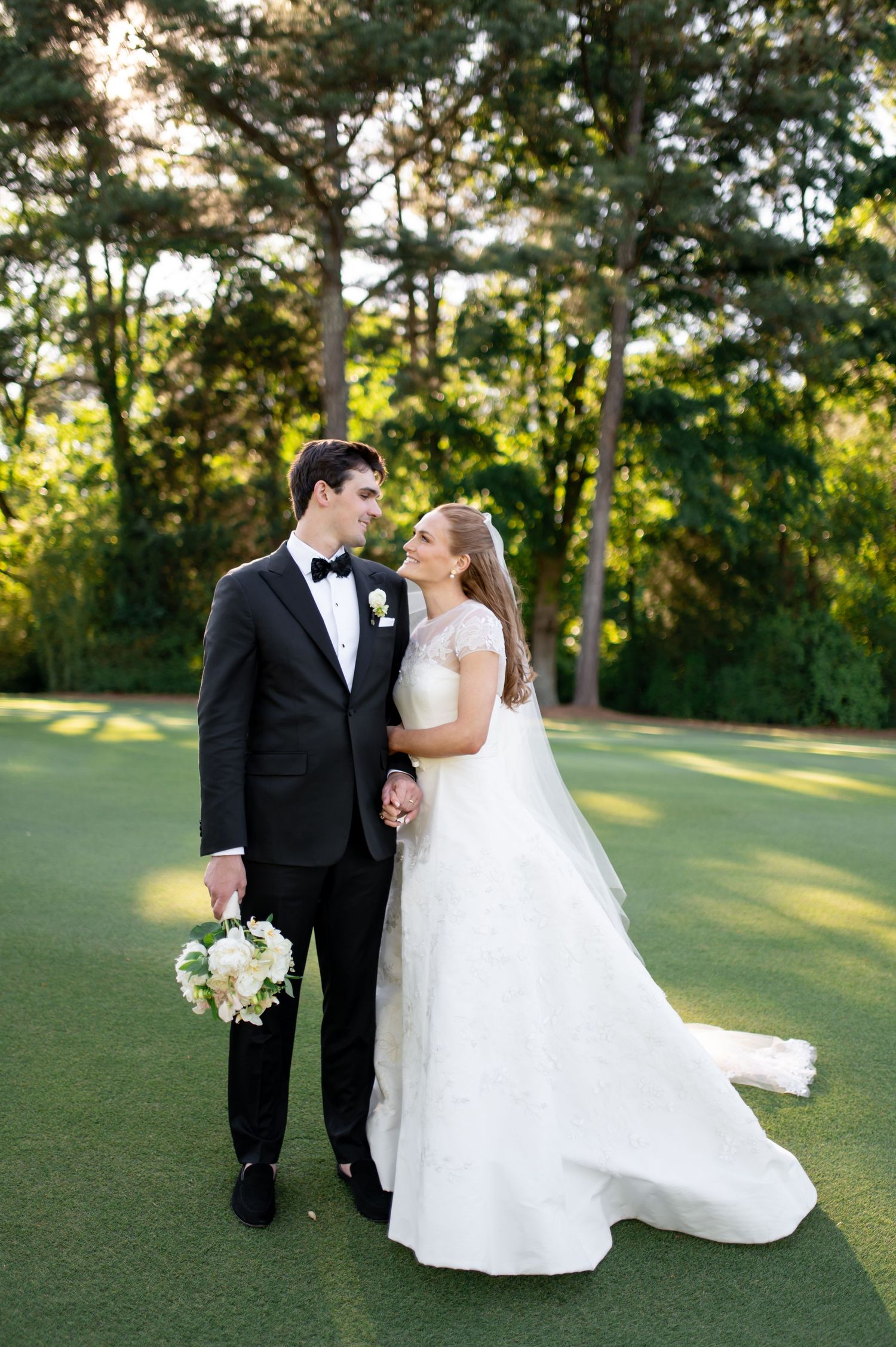 Bride and groom gazing at each other on lush golf course with sunlit trees behind them.