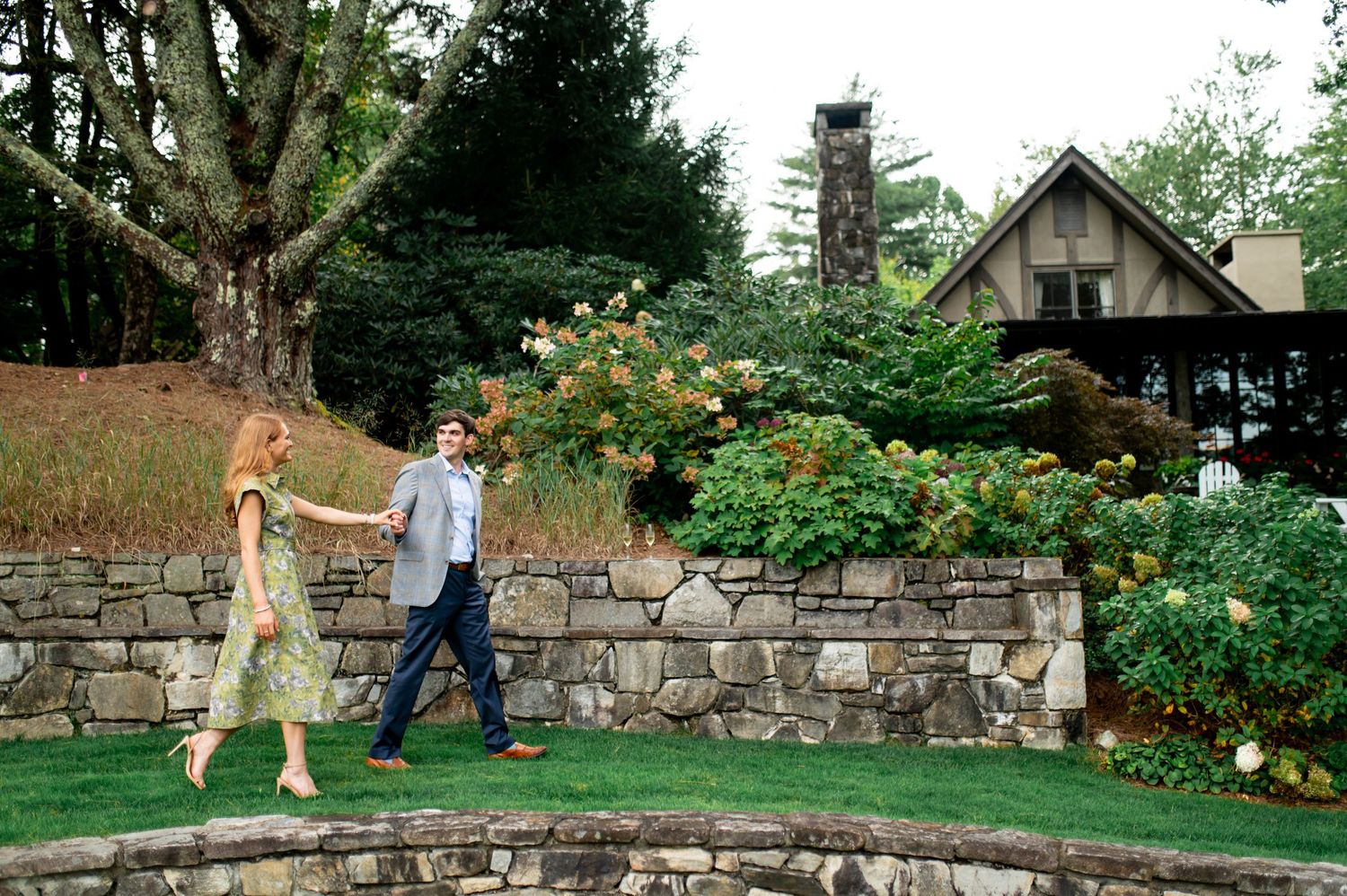 A couple walks along a rustic stone wall in a garden setting.