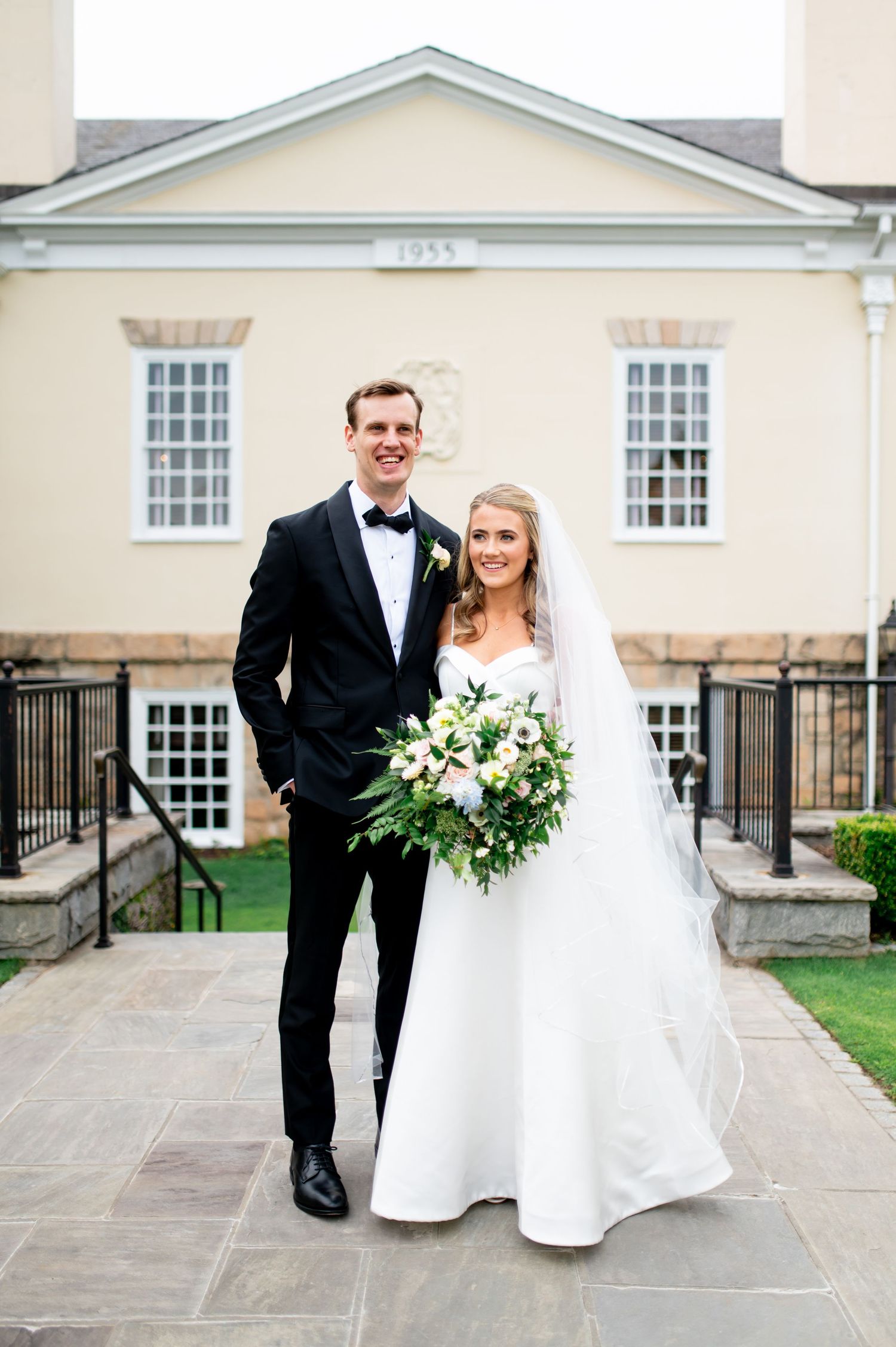 Elegant wedding couple poses in front of classic cream-colored building with architectural details.