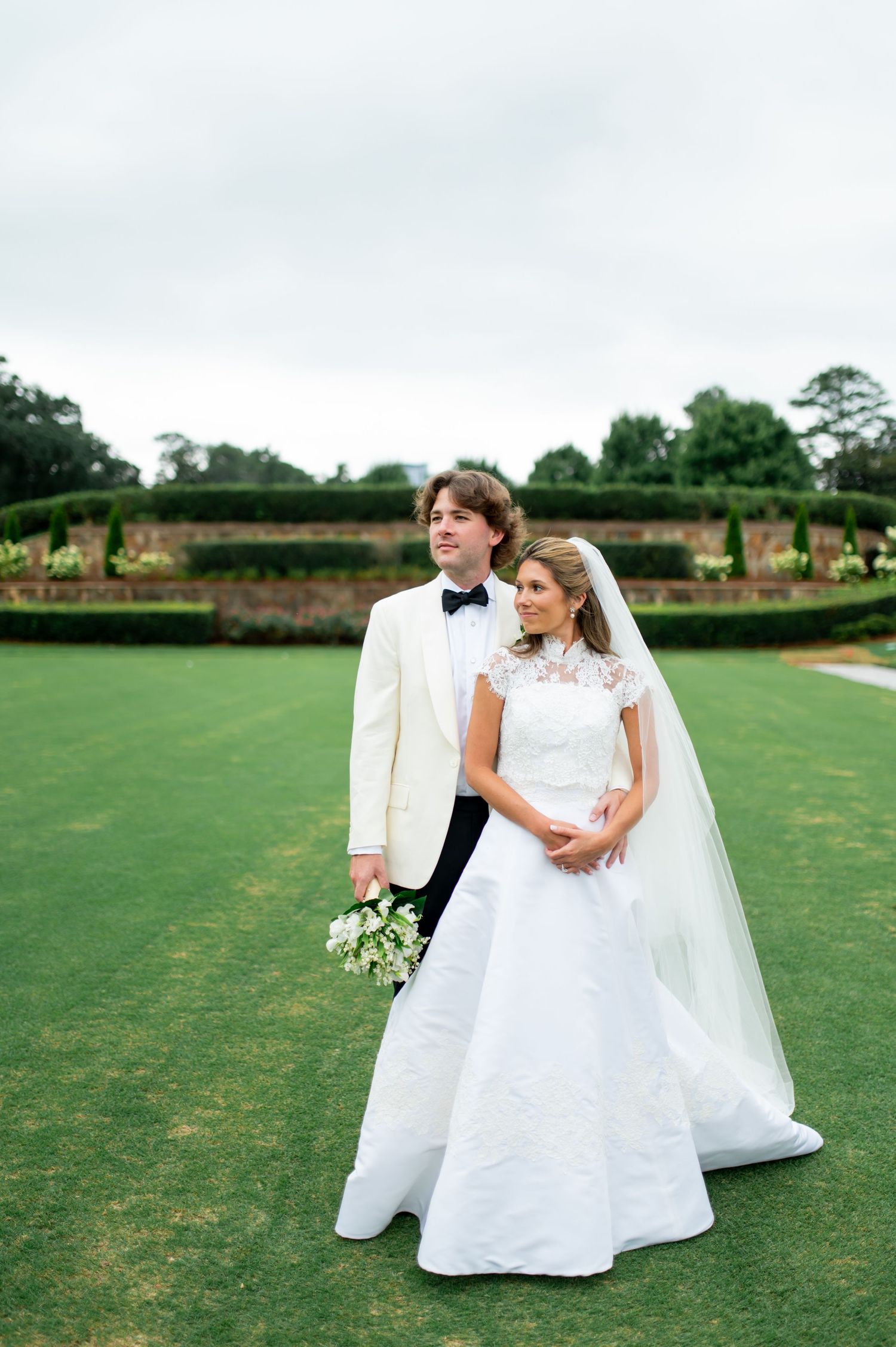 Elegant couple poses on a garden lawn in formal wedding attire with white flowers and flowing veil.
