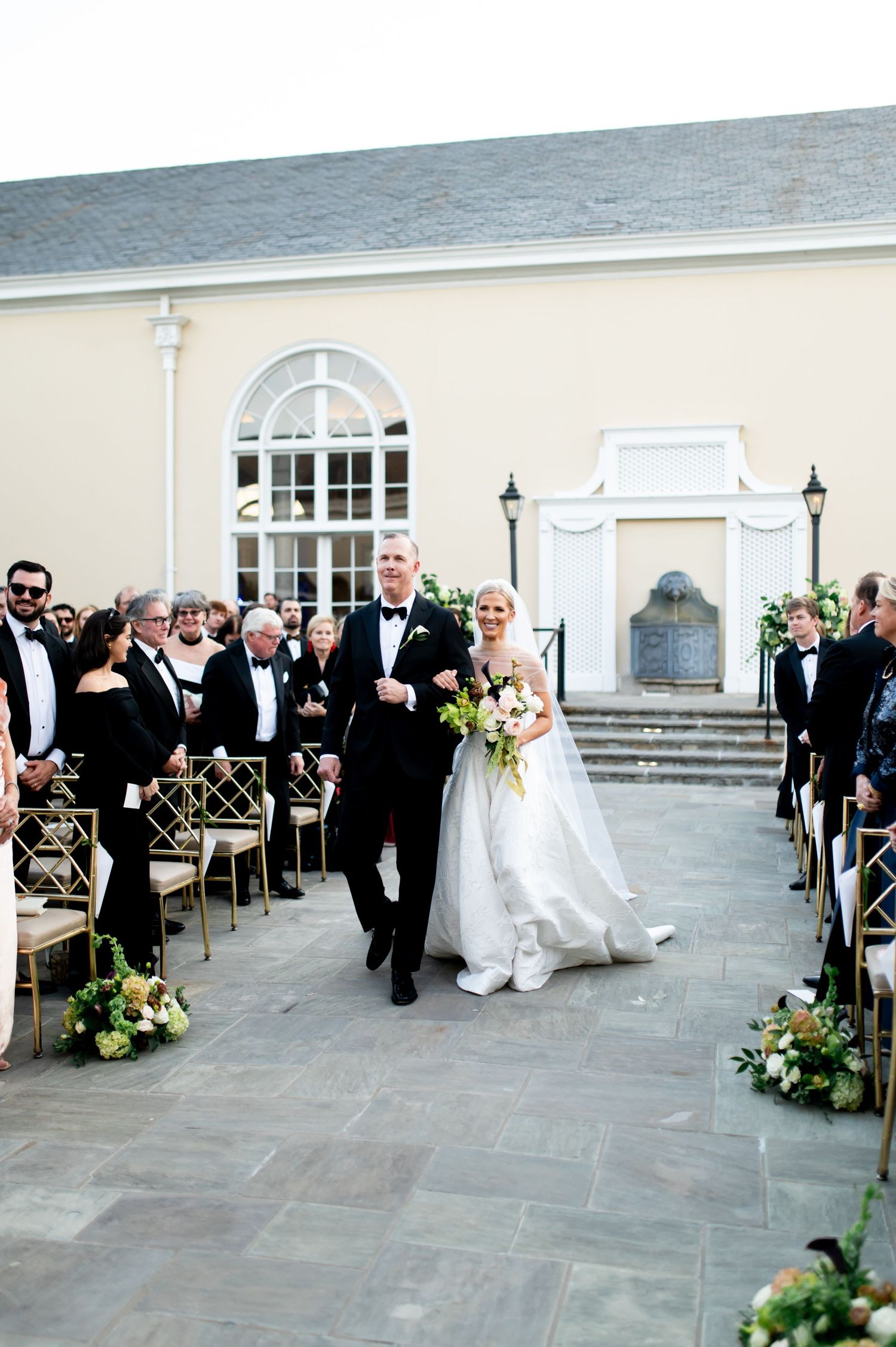 Wedding ceremony setup on stone patio with white columns and elegant architecture in background.