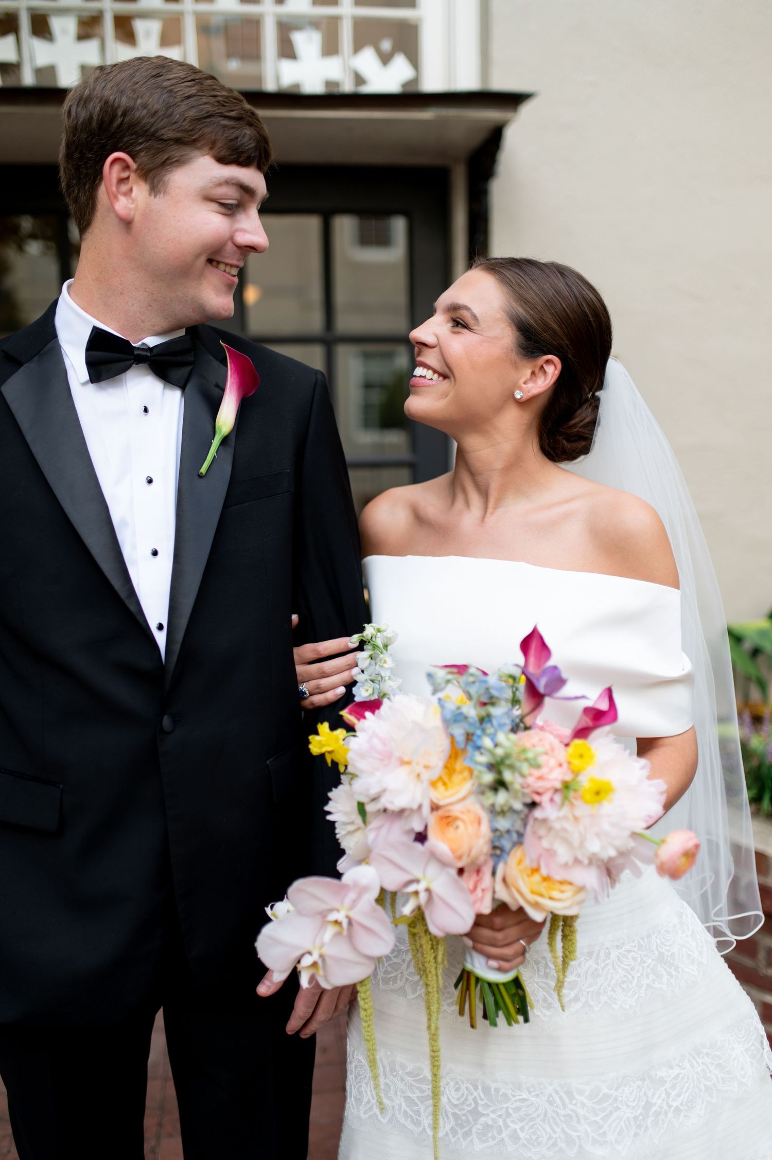 Bride and groom sharing a joyful moment, she holds a colorful bouquet in a white off-shoulder gown.