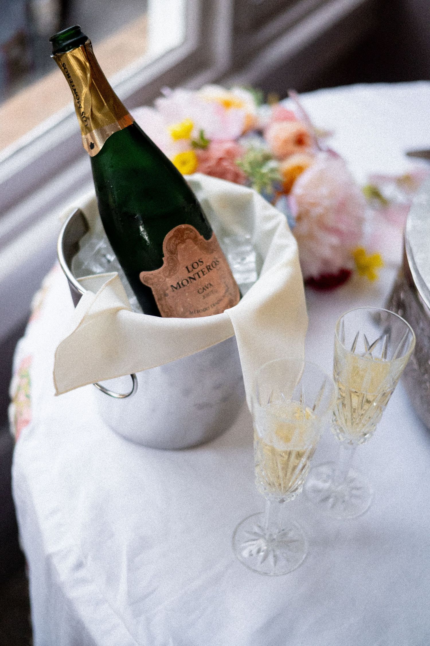 Champagne bottle chilling in ice bucket beside crystal flutes and floral arrangement on white linen.