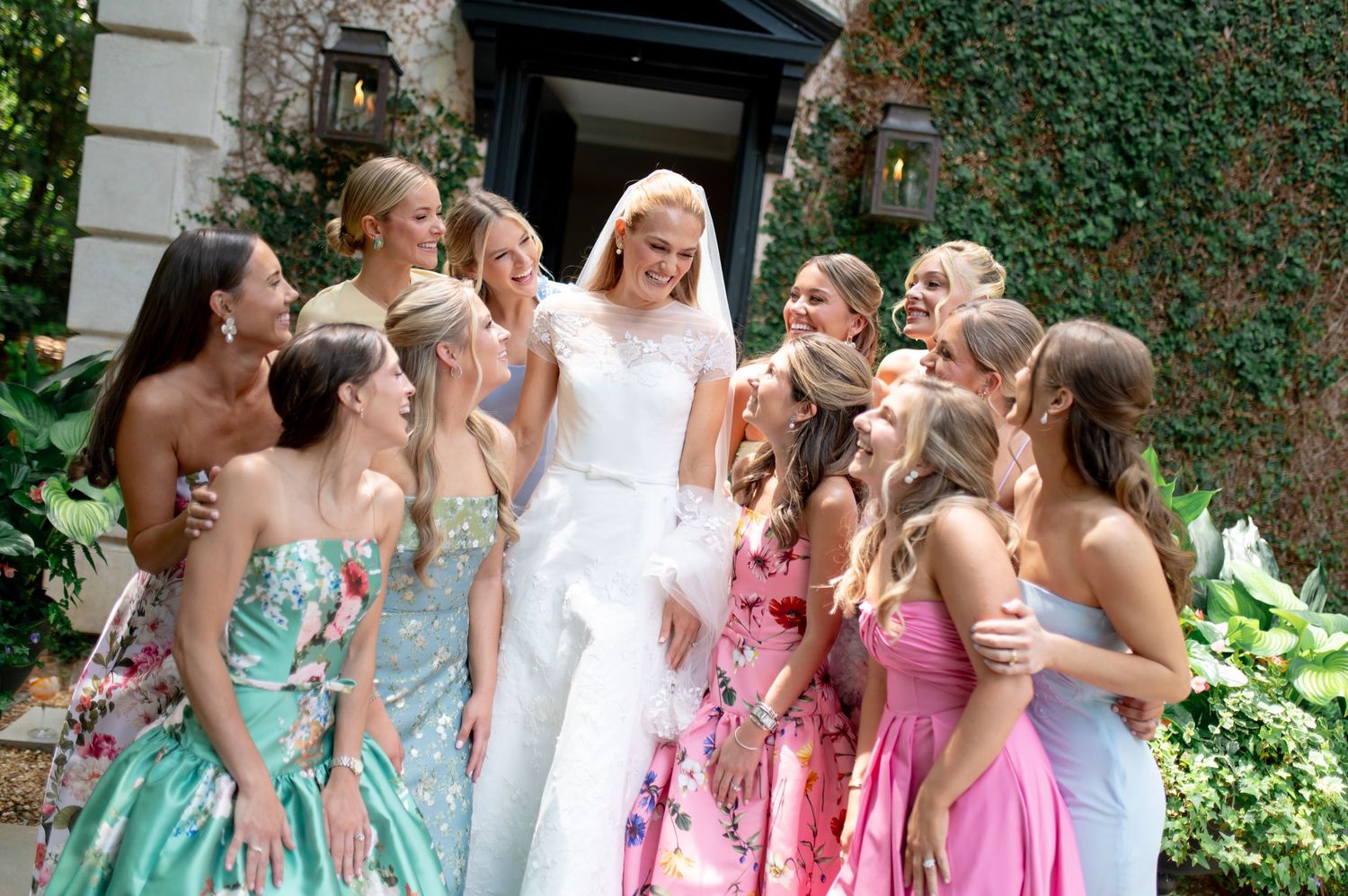Blonde bride in white gown surrounded by smiling bridesmaids in colorful floral dresses outside ivy-covered venue.