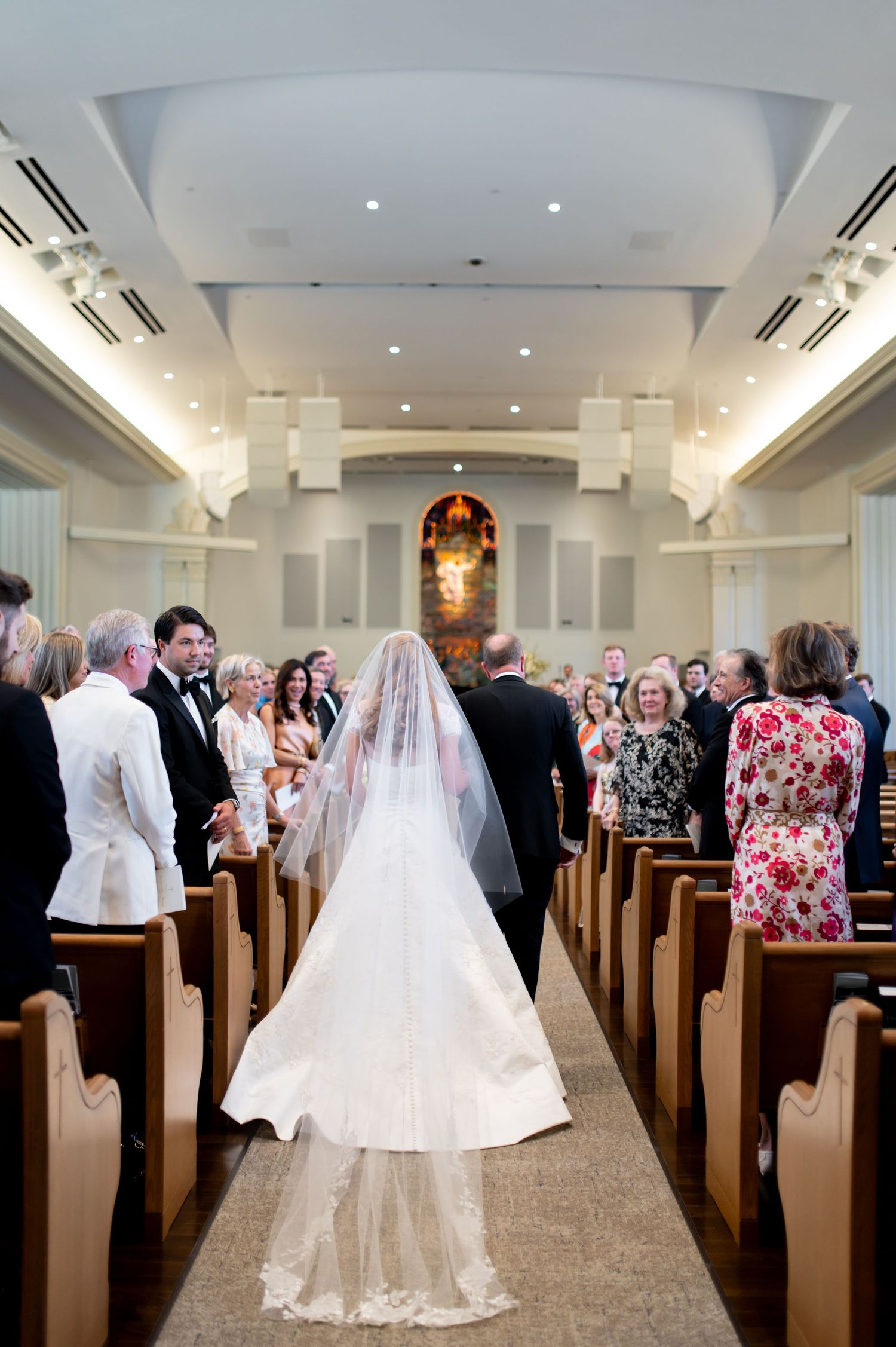Bride walks down church aisle in cathedral veil toward altar with seated guests.