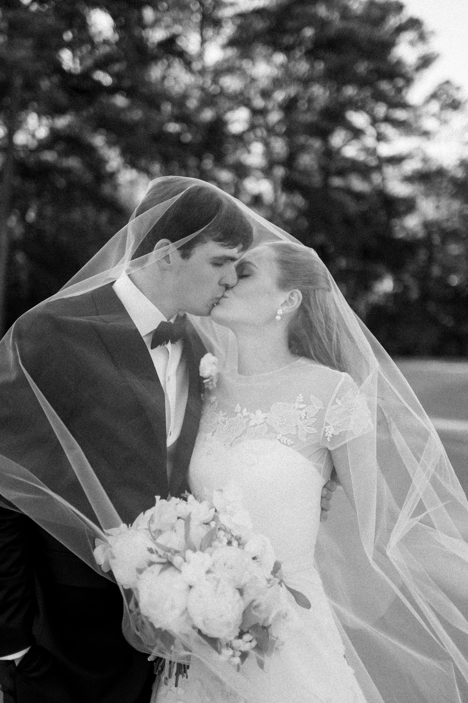 Bride and groom share romantic kiss under flowing veil in black and white wedding portrait.