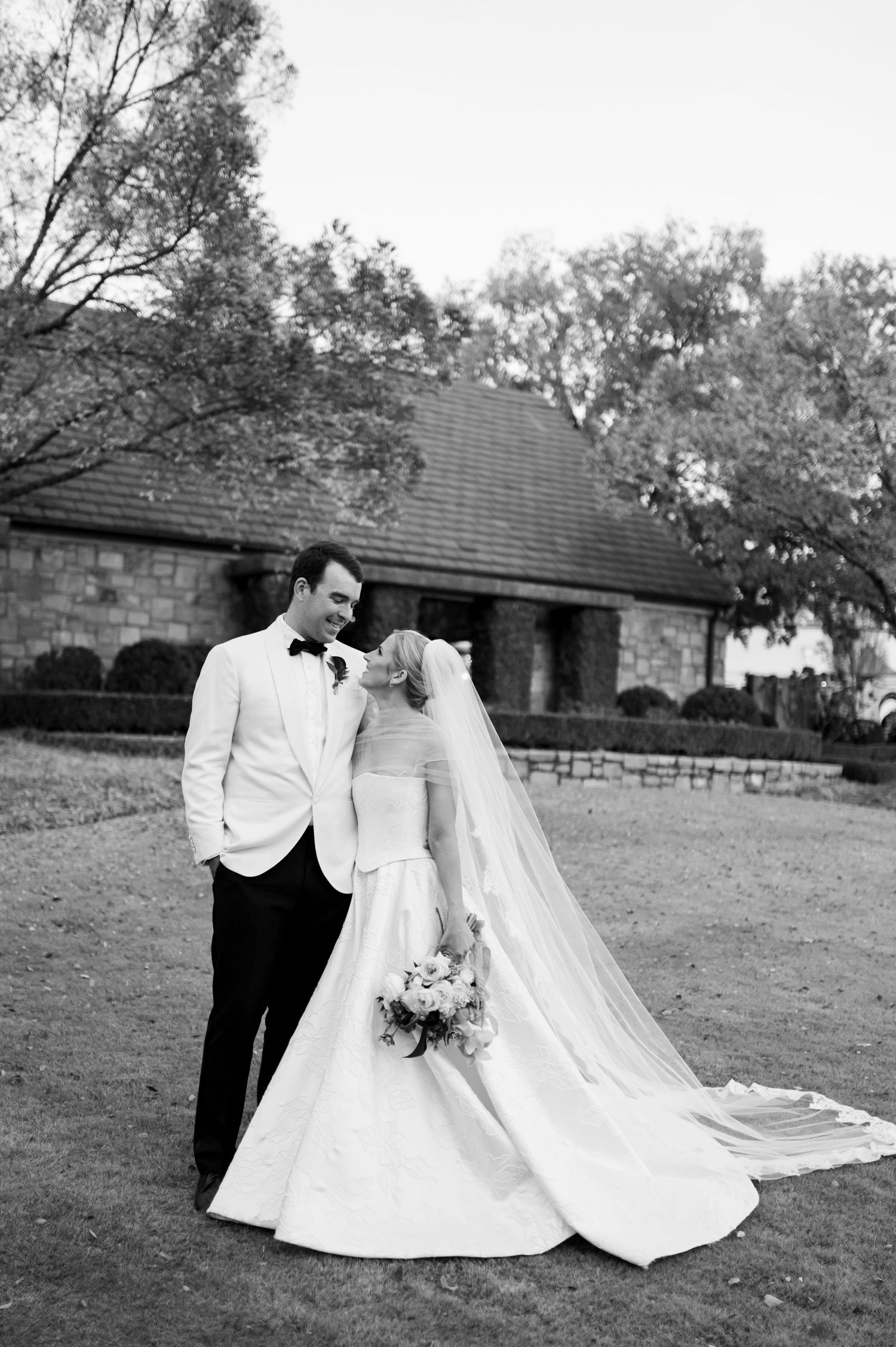 A bride and groom in classic white and black attire pose together in a rustic outdoor setting.