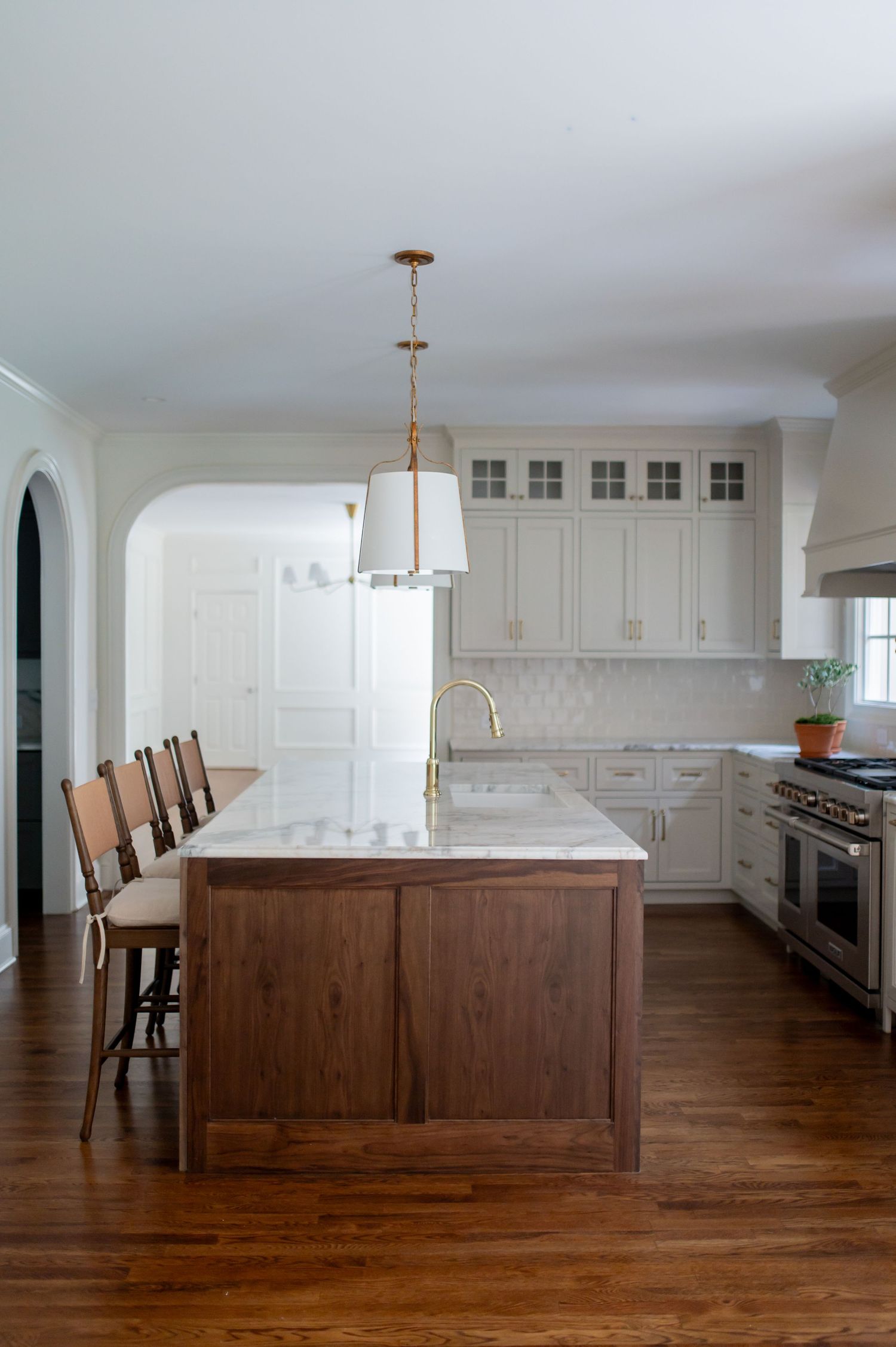 White kitchen with walnut island, marble countertop, brass pendant light, bar stools, and stainless steel range on hardwood floors.