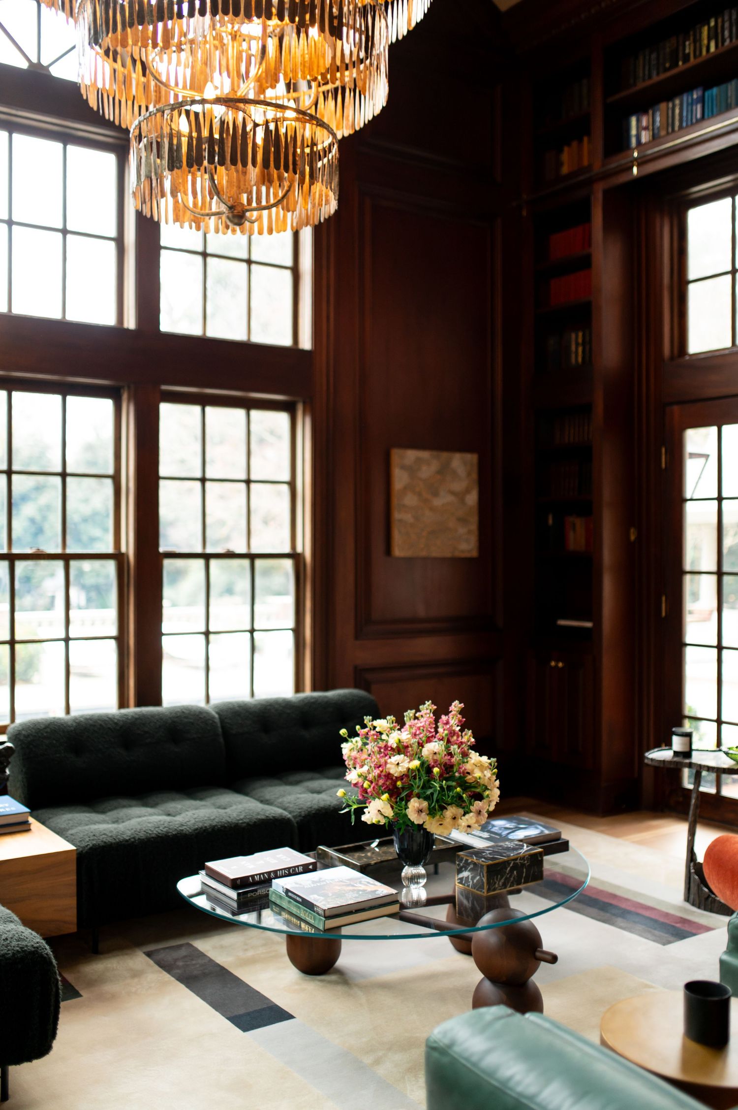 Elegant library living room with dark wood paneling, tufted sofa, glass coffee table, floral arrangement, and gold chandelier.