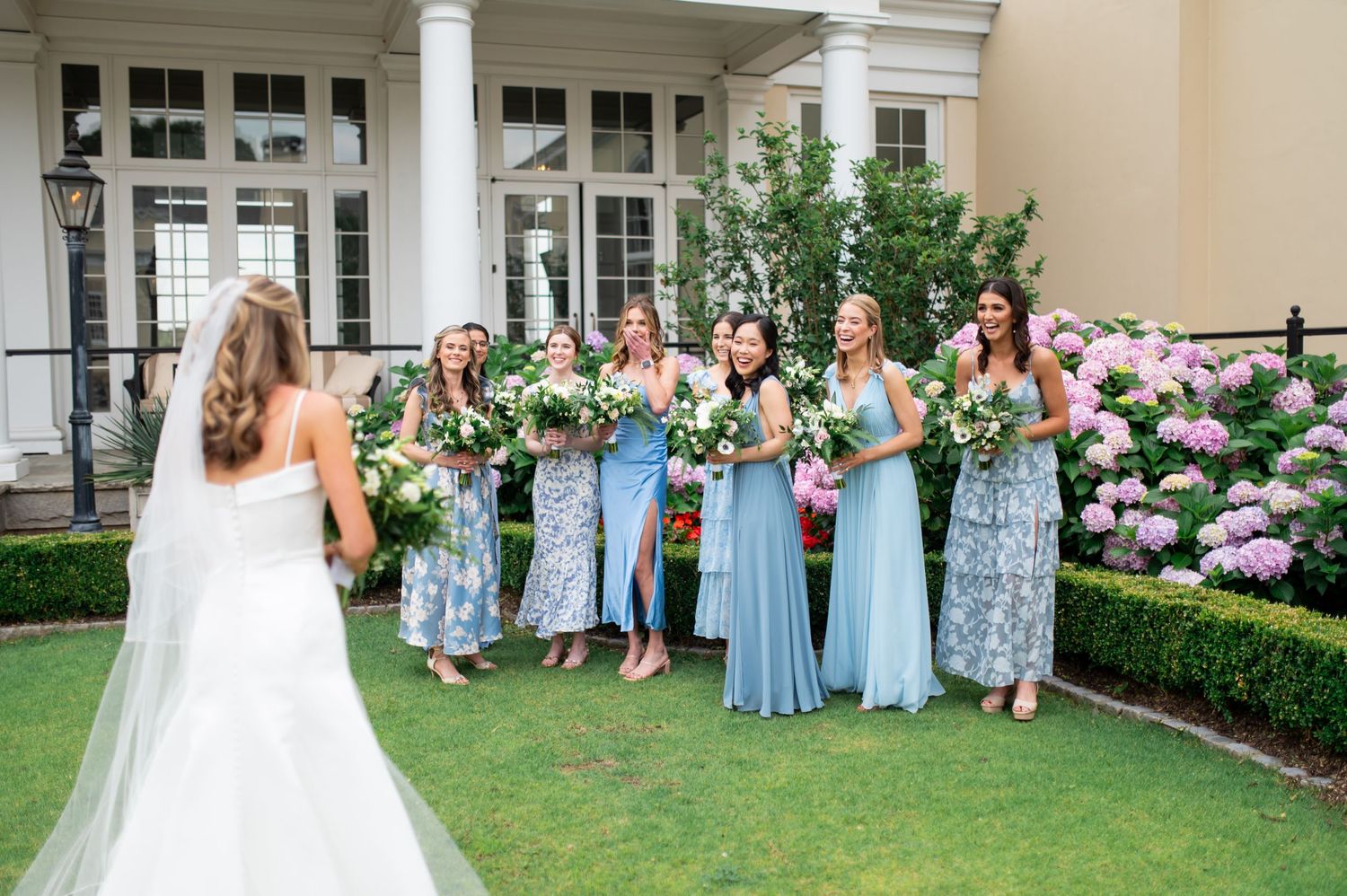 Bridal party in blue dresses stands on lawn while bride poses with bouquet.