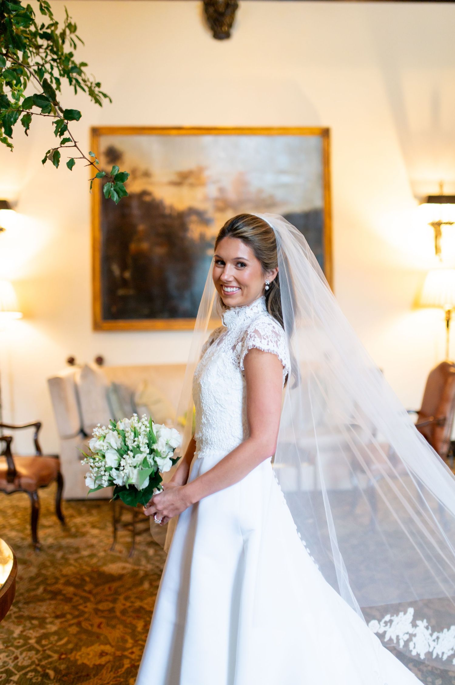 Smiling bride in lace wedding gown and veil holds white bouquet indoors before elegant painting backdrop.