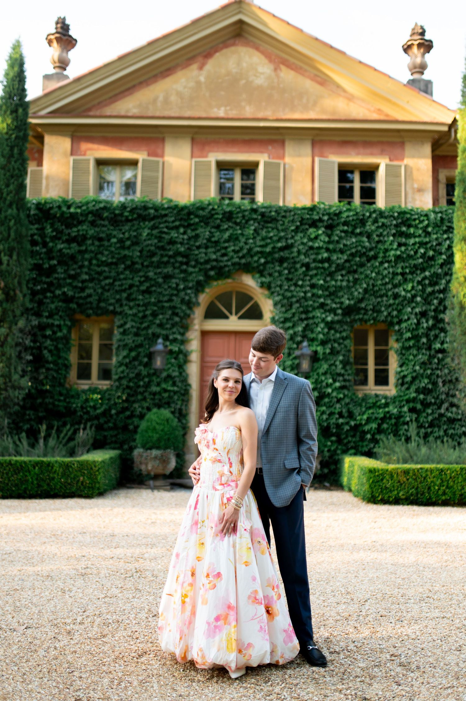 Couple posing in front of ivy-covered Italian villa, woman in floral gown and man in gray suit on gravel path.
