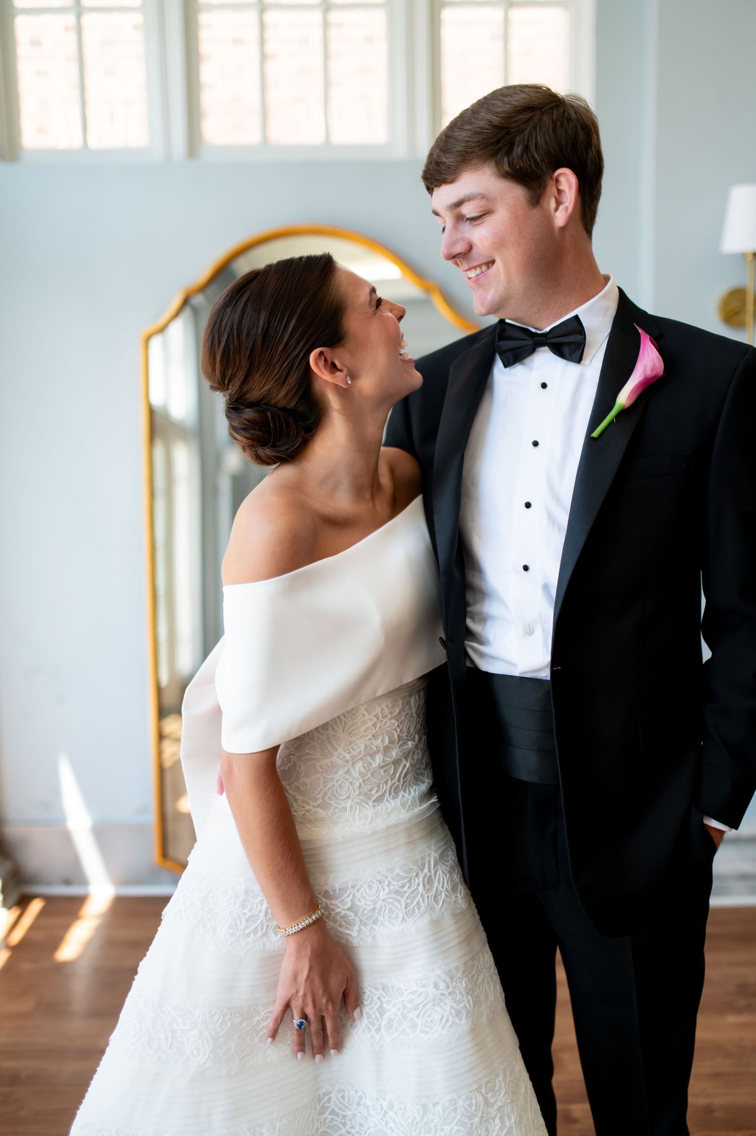 Bride in off-shoulder white gown and groom in black tuxedo with pink boutonniere share a romantic moment indoors.
