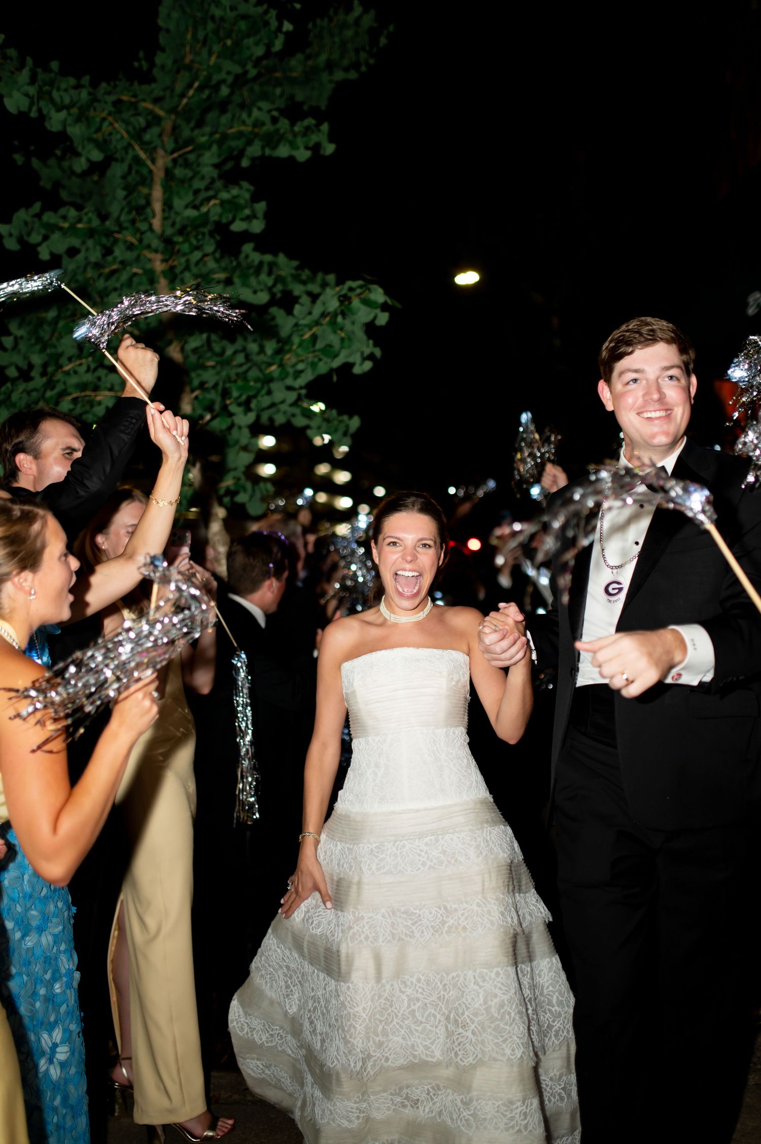 Smiling bride and groom walk through sparkler send-off tunnel with cheering guests at nighttime wedding exit.