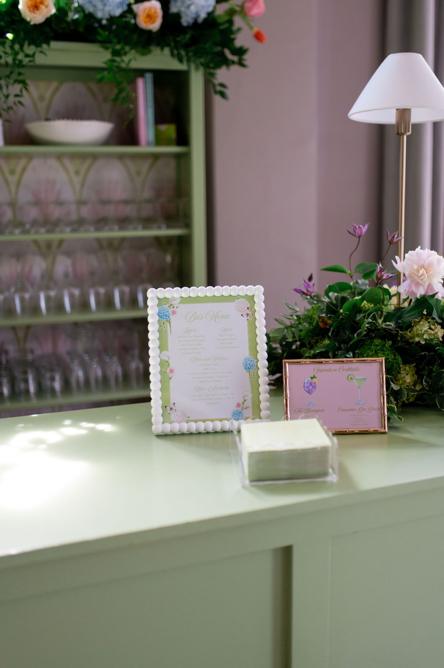 Elegant wedding reception table with floral menu display, pink frame, and green shelving backdrop.