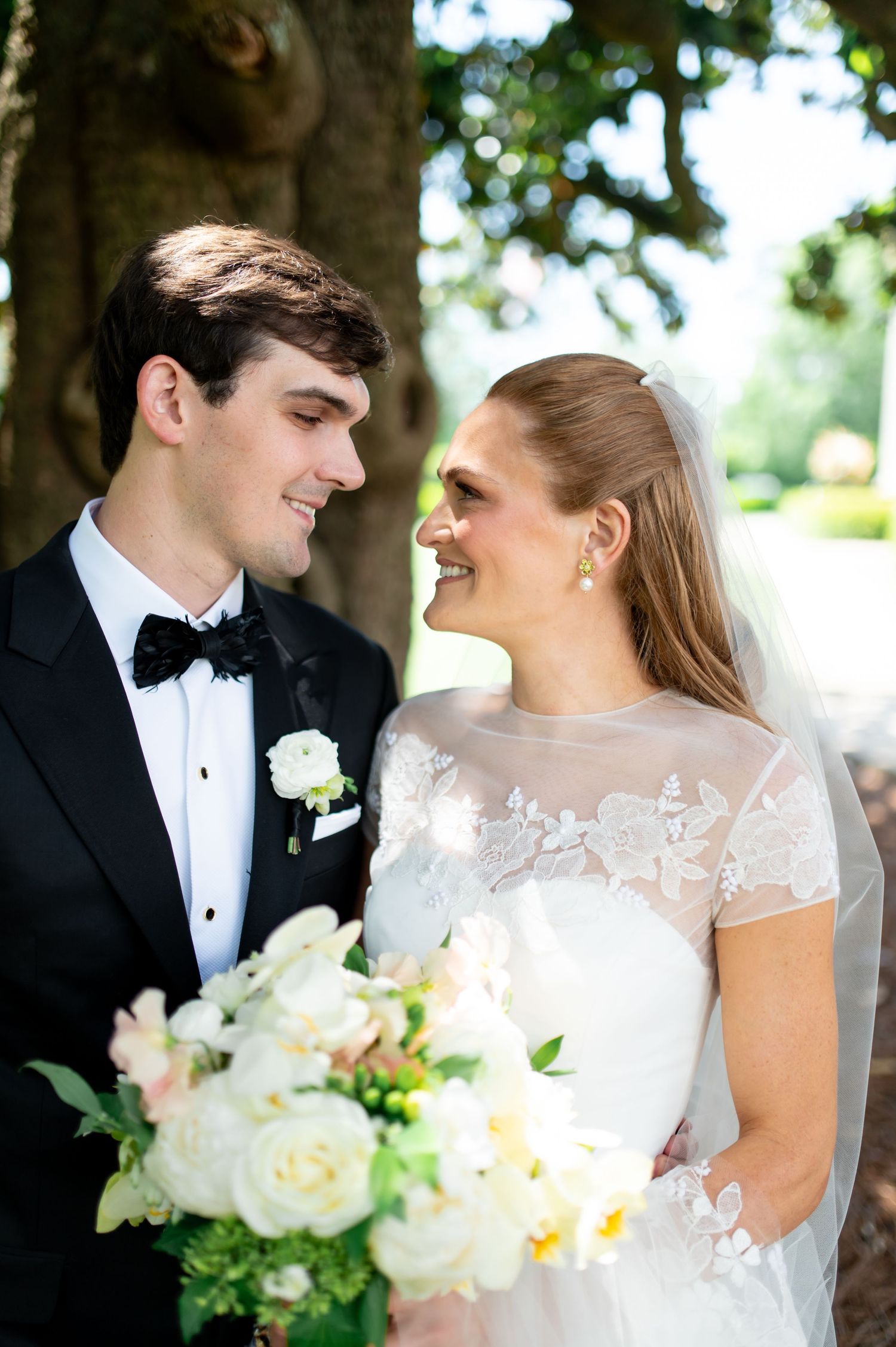Couple sharing a loving gaze, bride in lace gown holding white bouquet under oak tree.