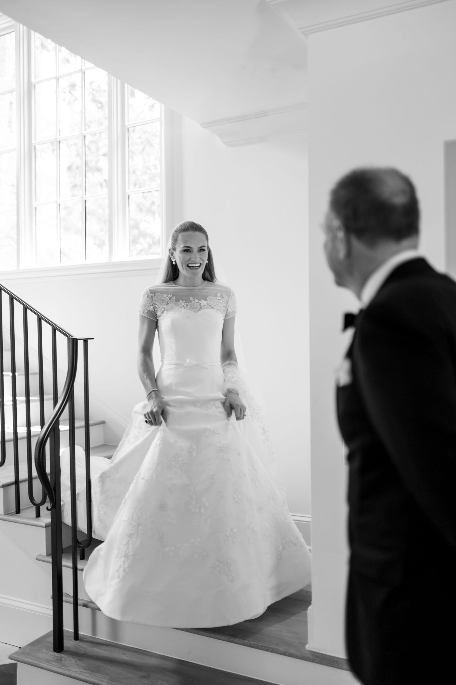 Bride in lace wedding gown smiling on staircase during first look with groom in tuxedo, black and white photo.