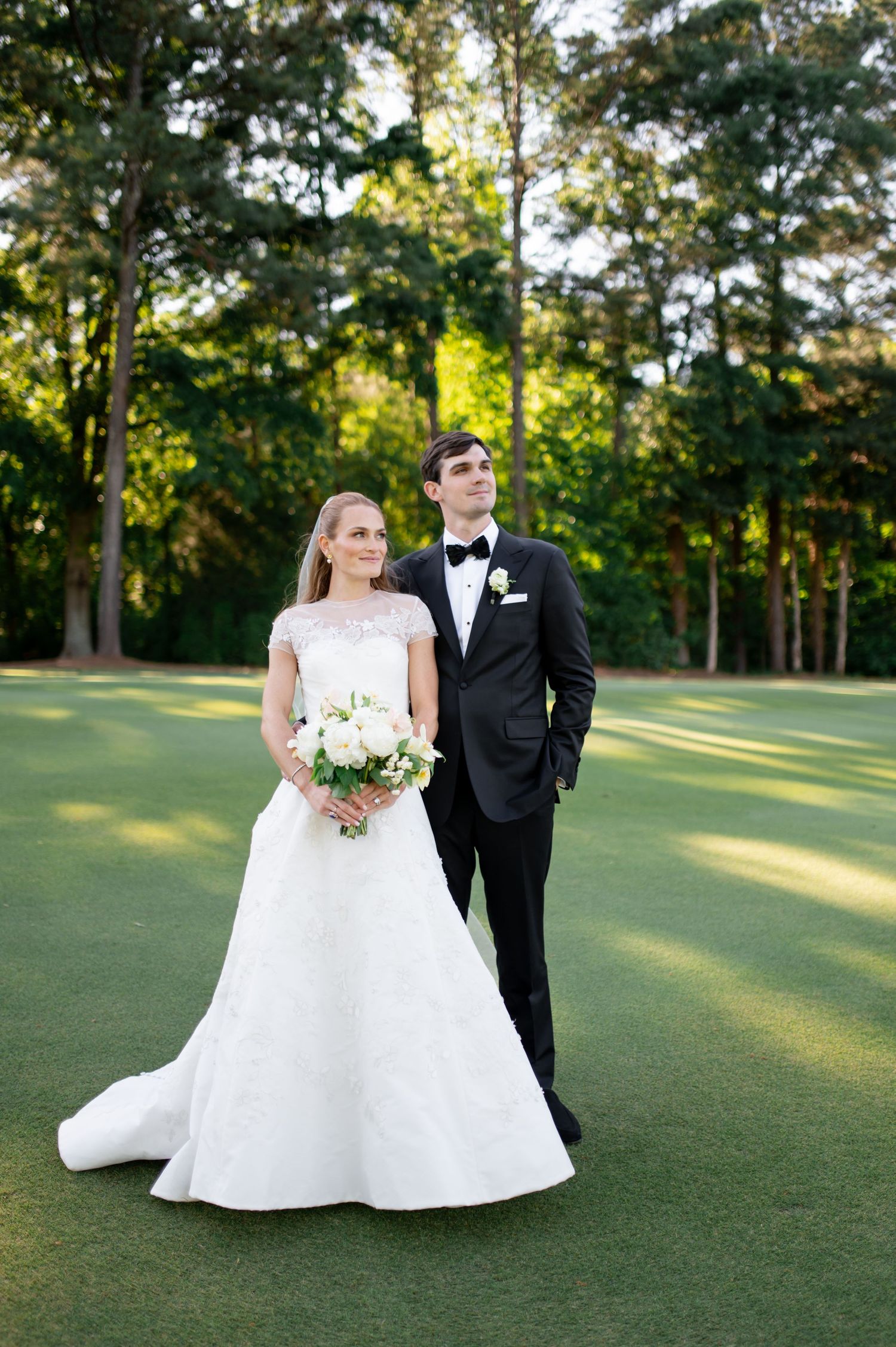 Bride and groom portrait on golf course green surrounded by tall pine trees at sunset.