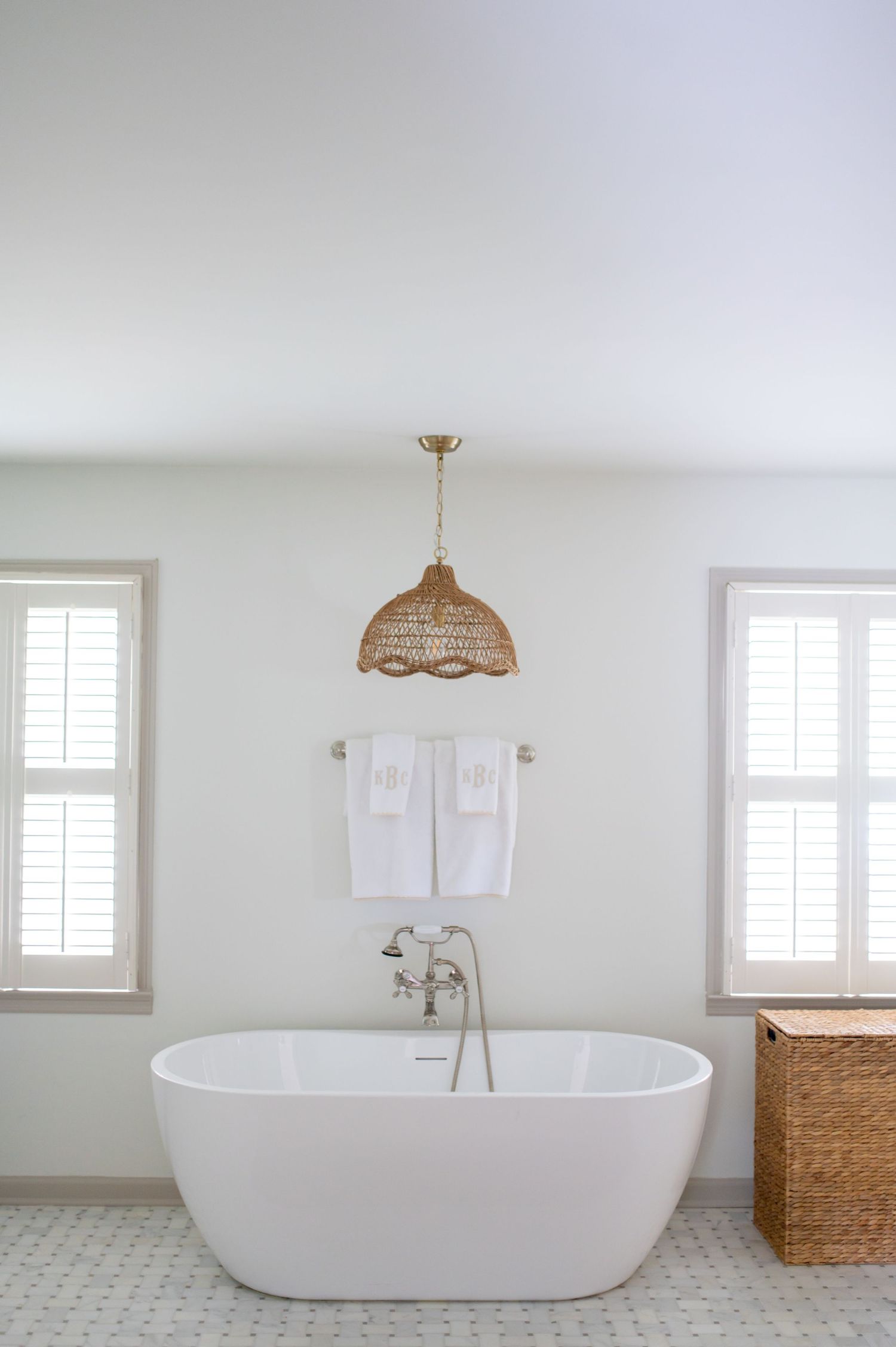 Modern freestanding white bathtub in minimalist bathroom with woven pendant light and white towels on wall.