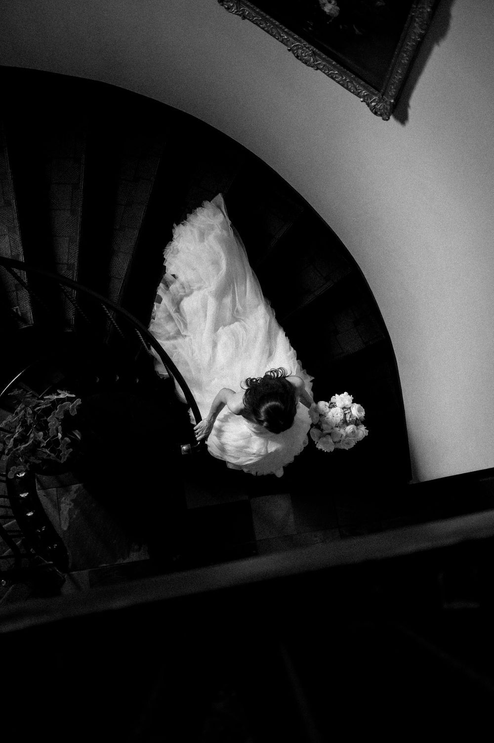 Black and white photo of a bride in a white gown descending a spiral staircase holding a bouquet.