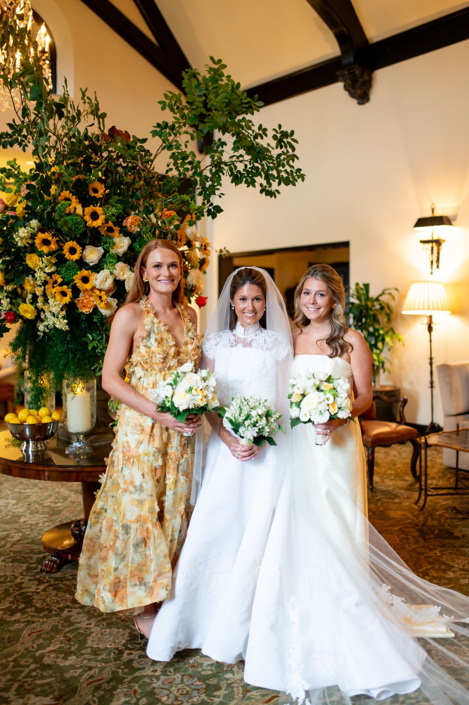 A bridal party poses in white dresses and yellow floral prints against an elegant indoor backdrop.