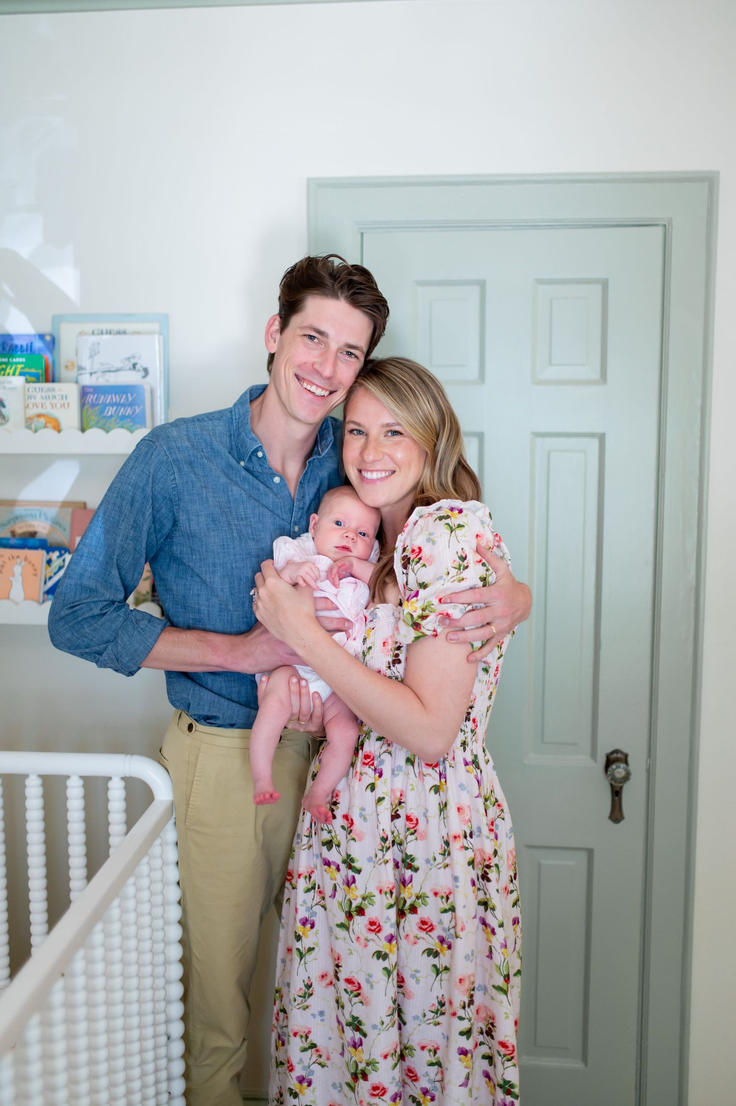 A couple holds their newborn baby in a white nursery with a crib and mint green door.