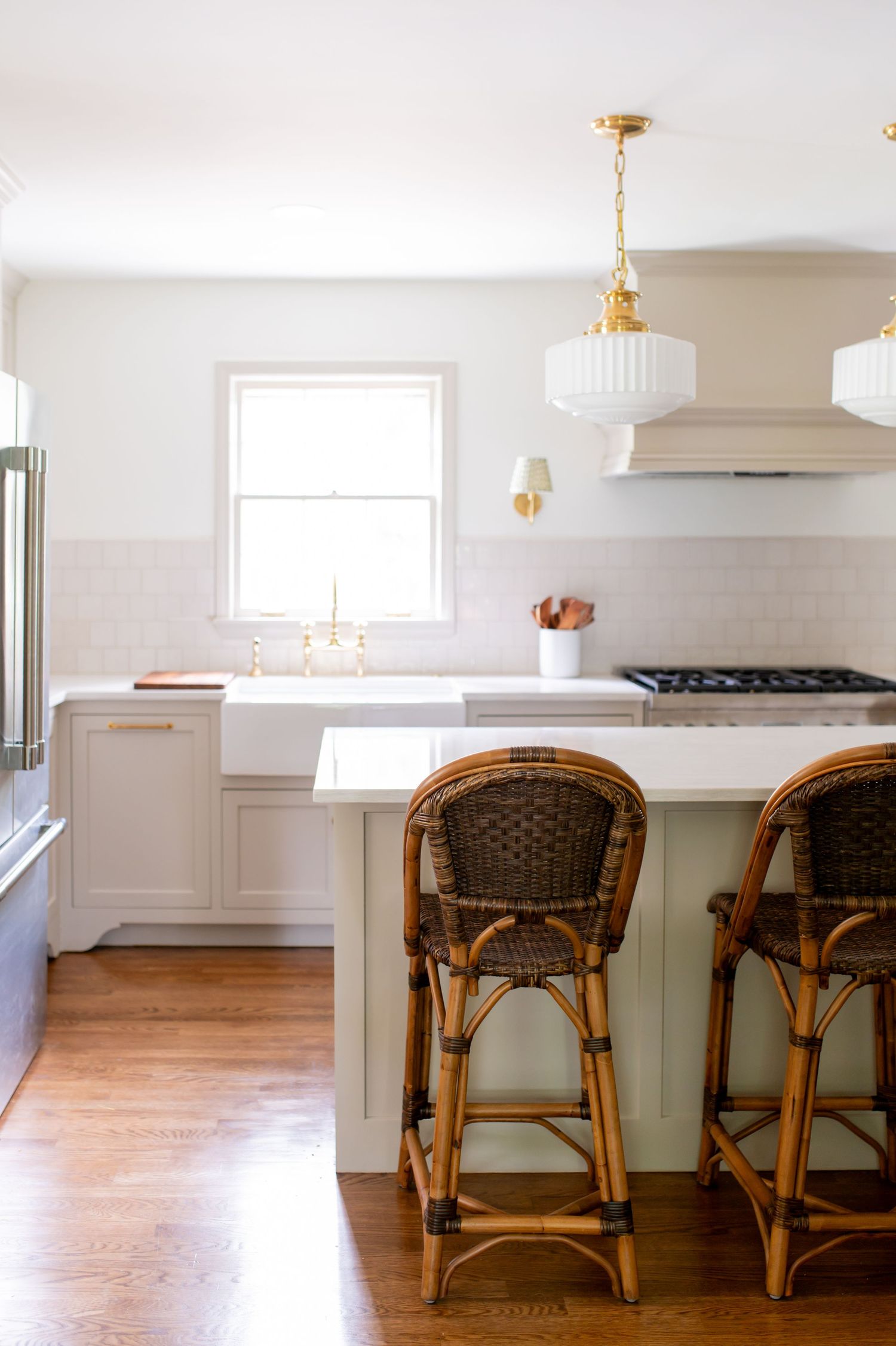 Modern kitchen with rattan bar stools, white cabinetry, and hardwood floors illuminated by gold pendant lights.