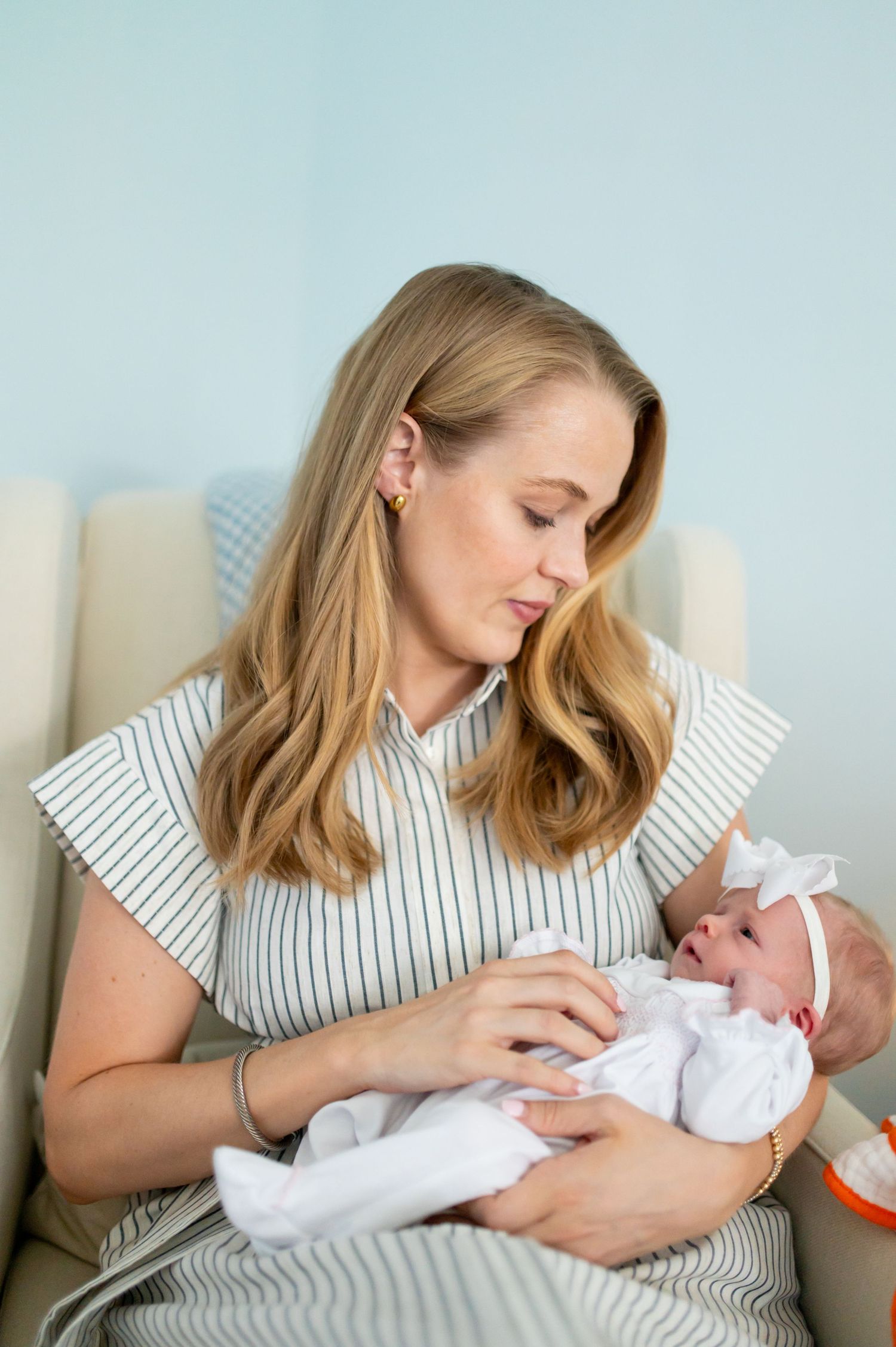 Someone in a striped dress tenderly holds a newborn baby wearing white on a cream colored chair.
