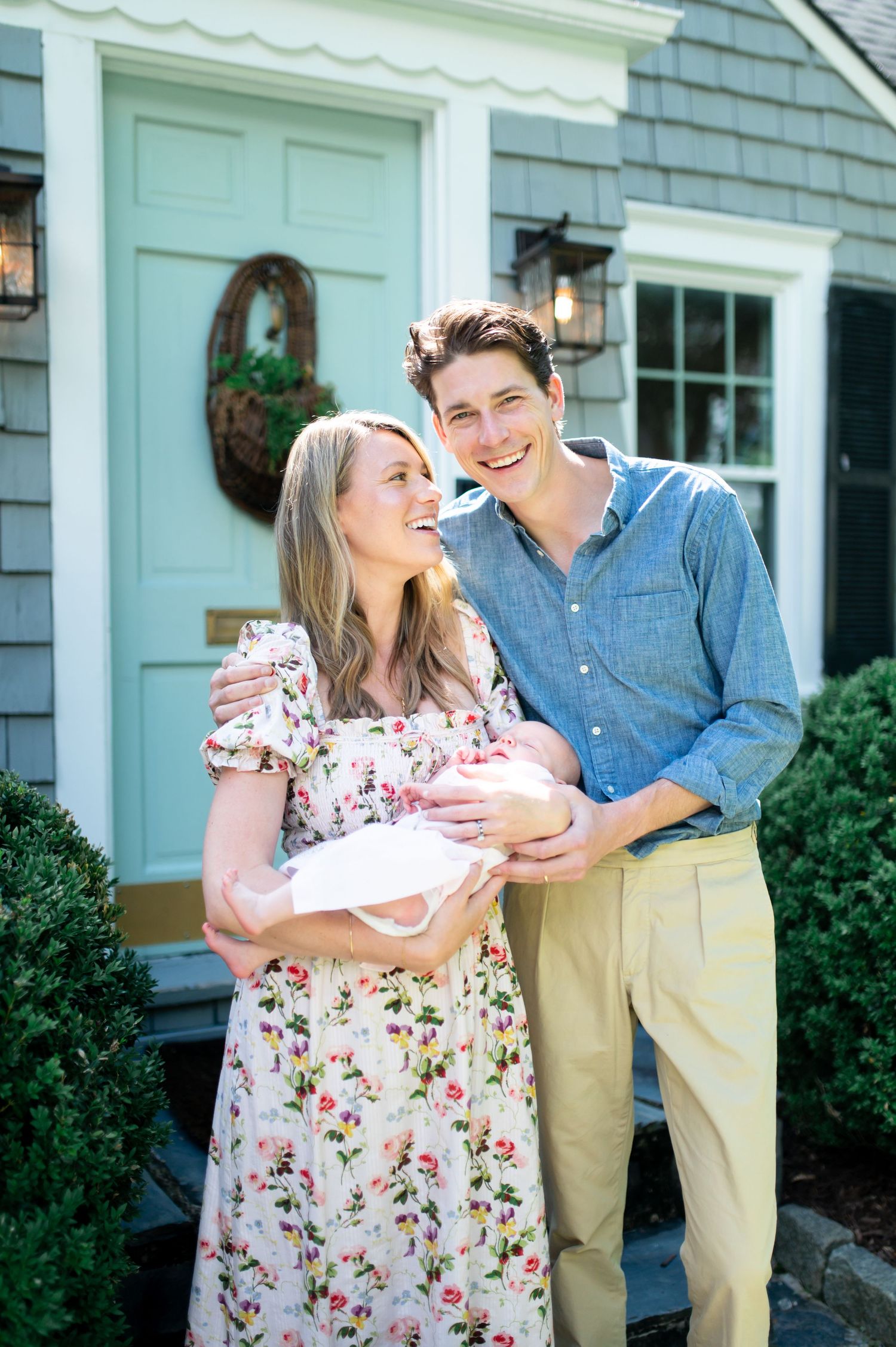 A couple shares a joyful moment while holding their newborn baby in front of a blue door and gray house.