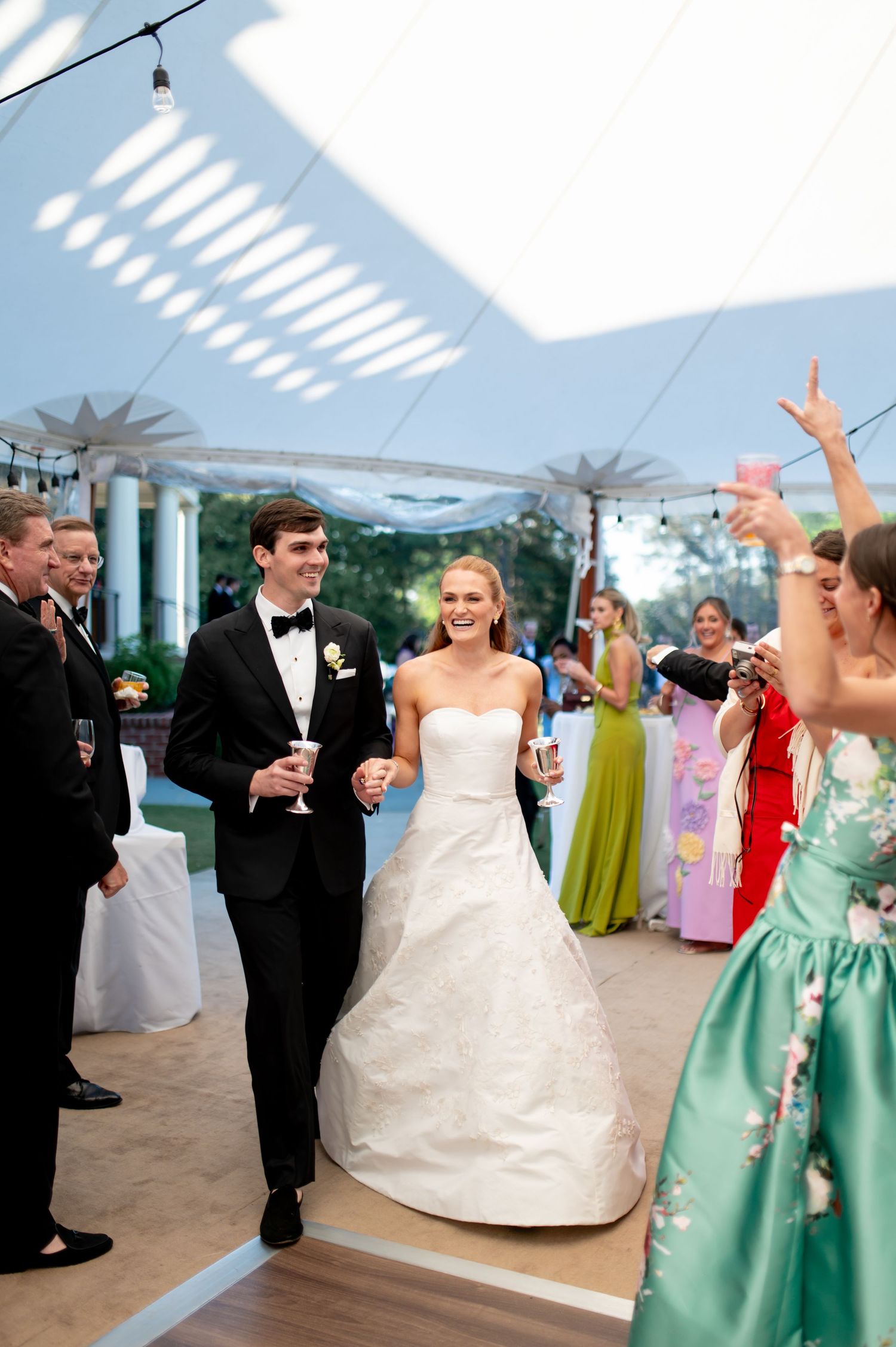 Bride and groom walk hand-in-hand at outdoor tented wedding reception, guests cheering in colorful formal attire.