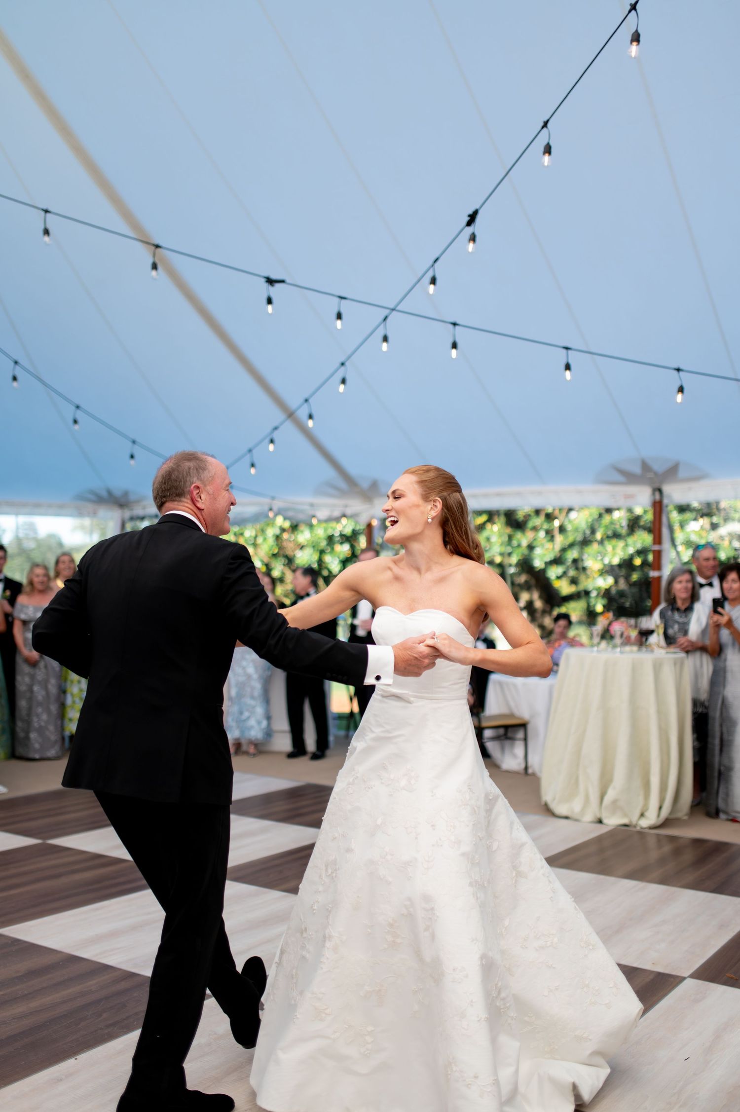 Bride in lace gown dances joyfully under string lights in elegant tent reception venue.
