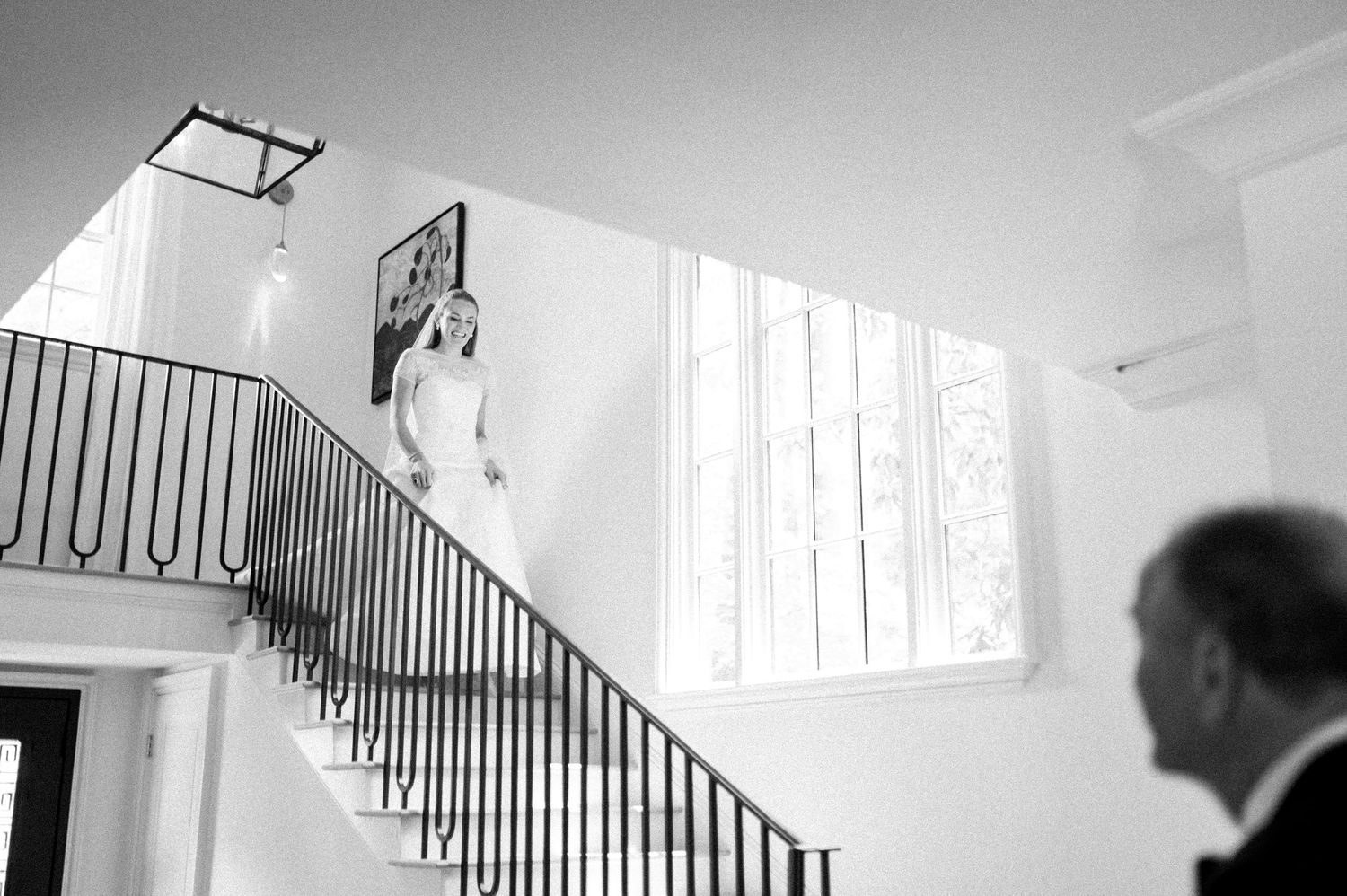 Bride in white gown descending elegant staircase in black and white wedding photo with guest in foreground.