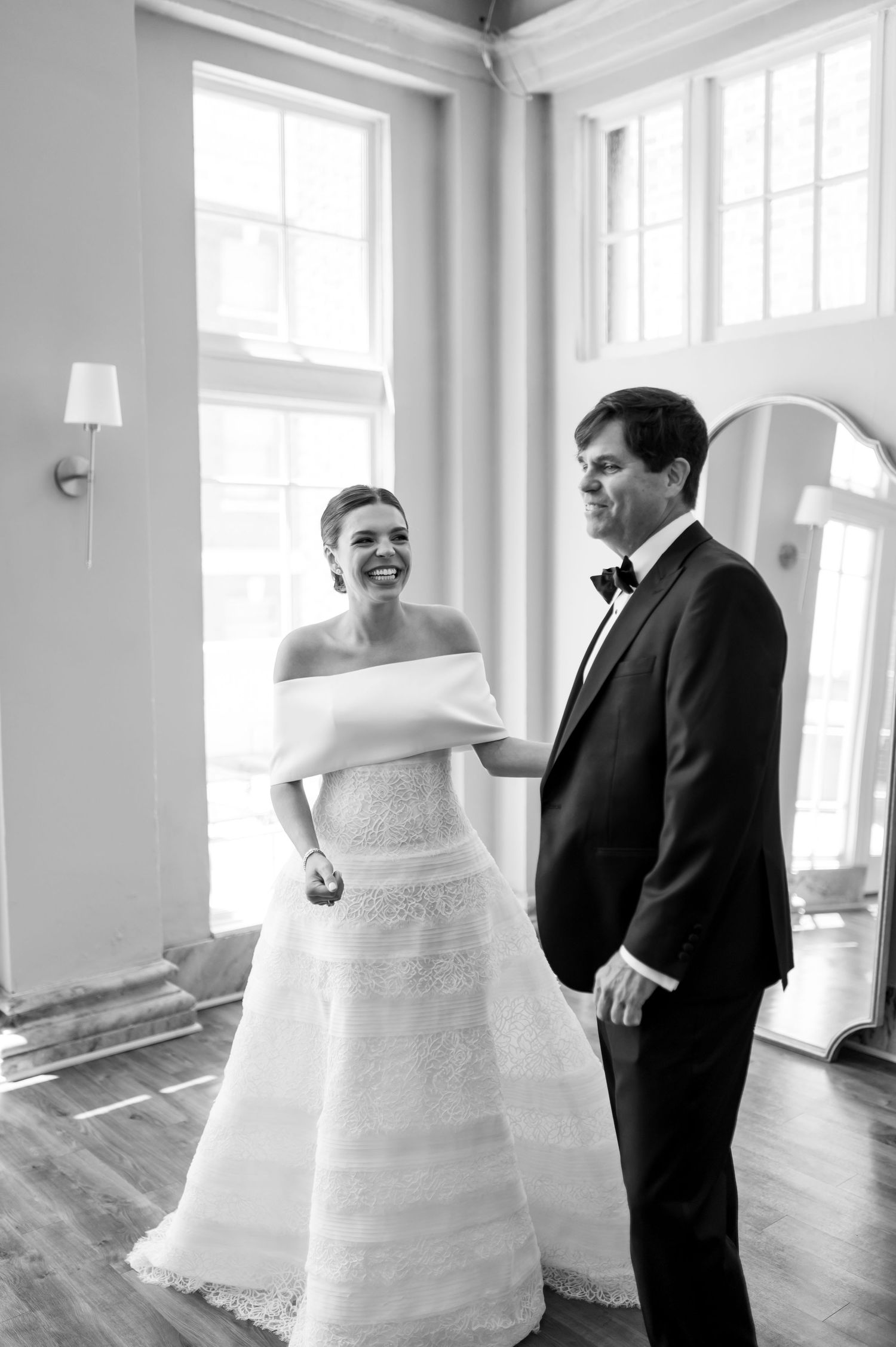 Black and white photo of a bride in an off-shoulder gown smiling at groom in tuxedo during first look.