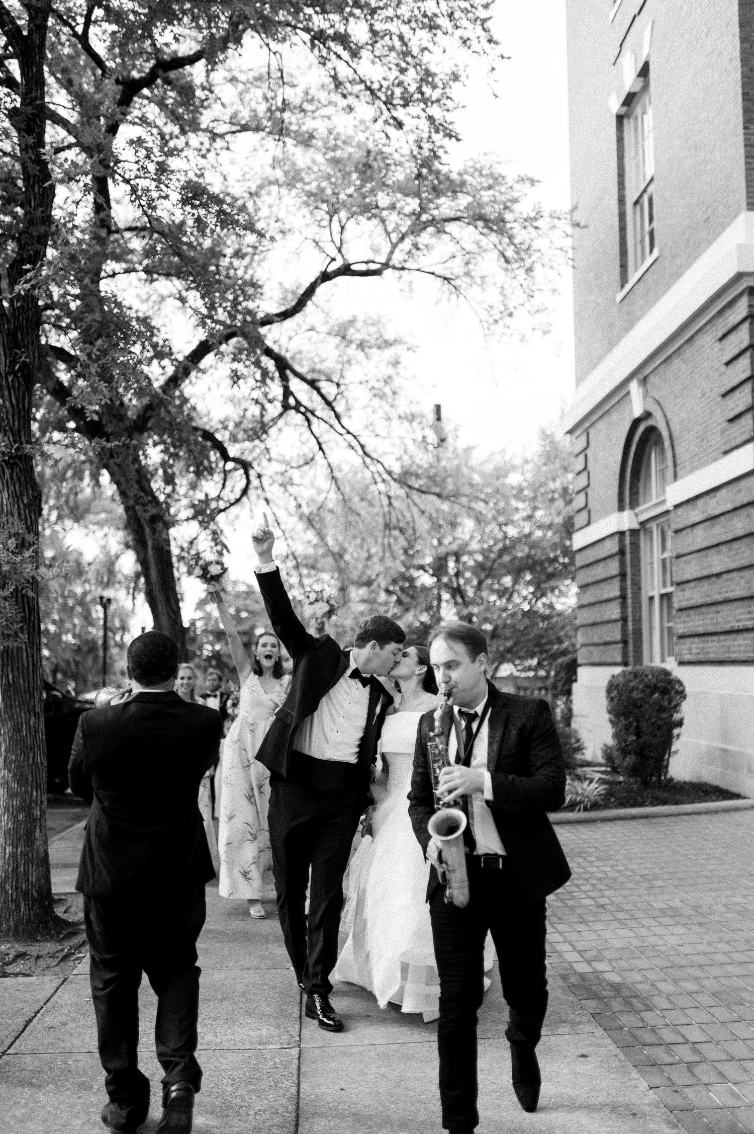 Black and white photo of newlyweds celebrating on sidewalk with saxophonist and guests cheering behind them.