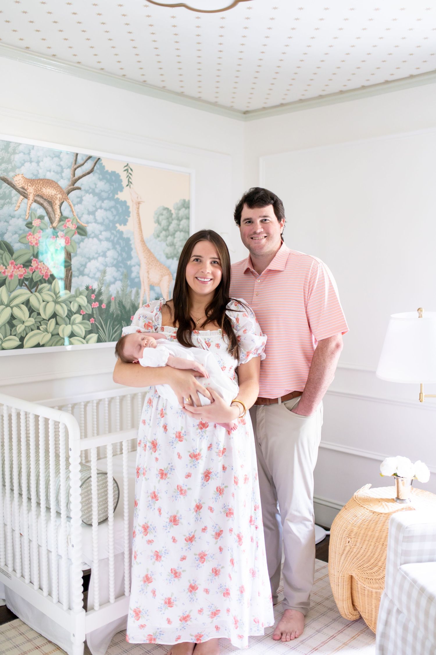 New parents smiling in a pastel nursery with white crib, floral mural, and mother holding newborn baby.