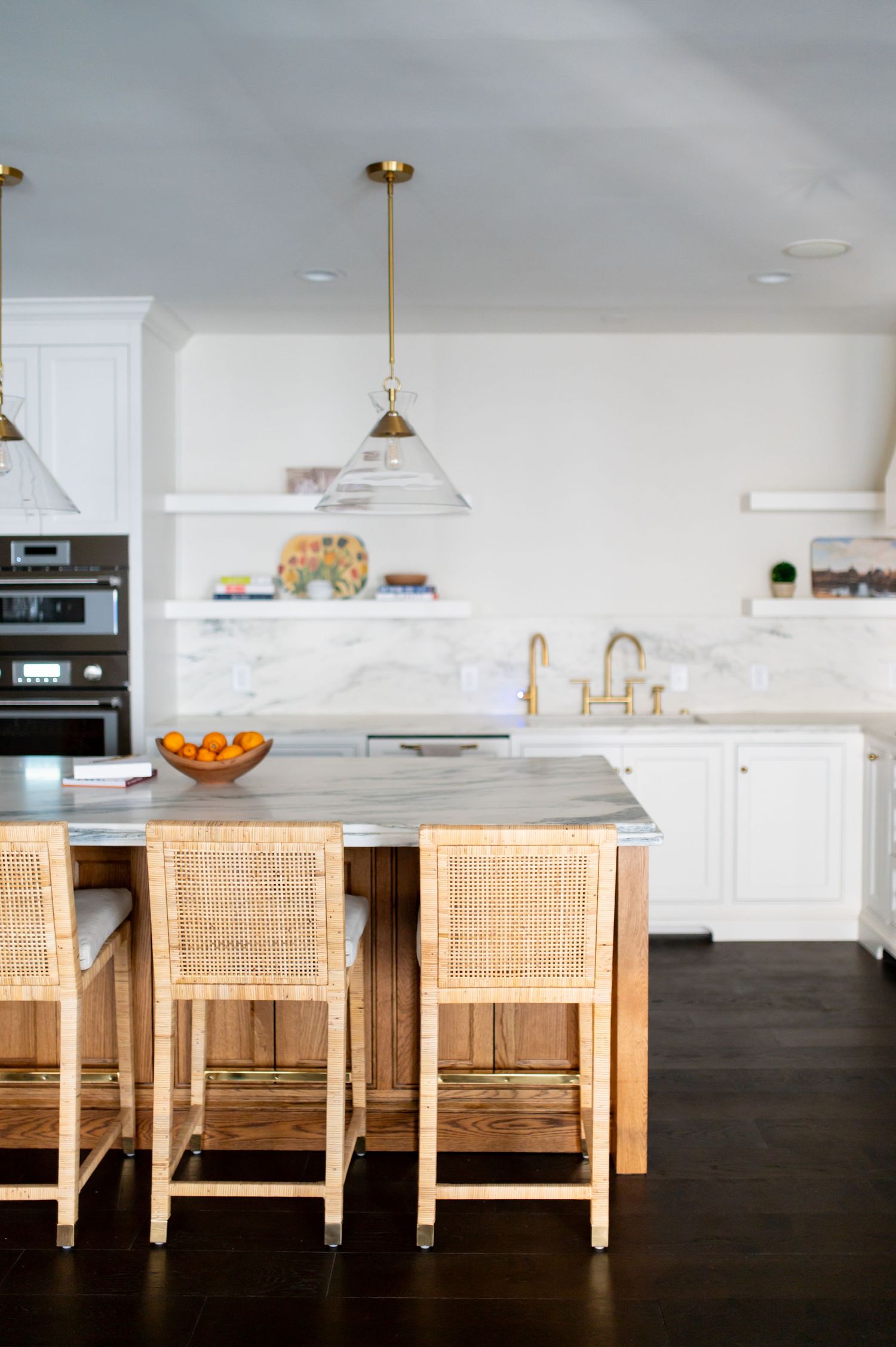 Modern white kitchen with rattan counter stools, marble countertops, brass fixtures and dark hardwood flooring.