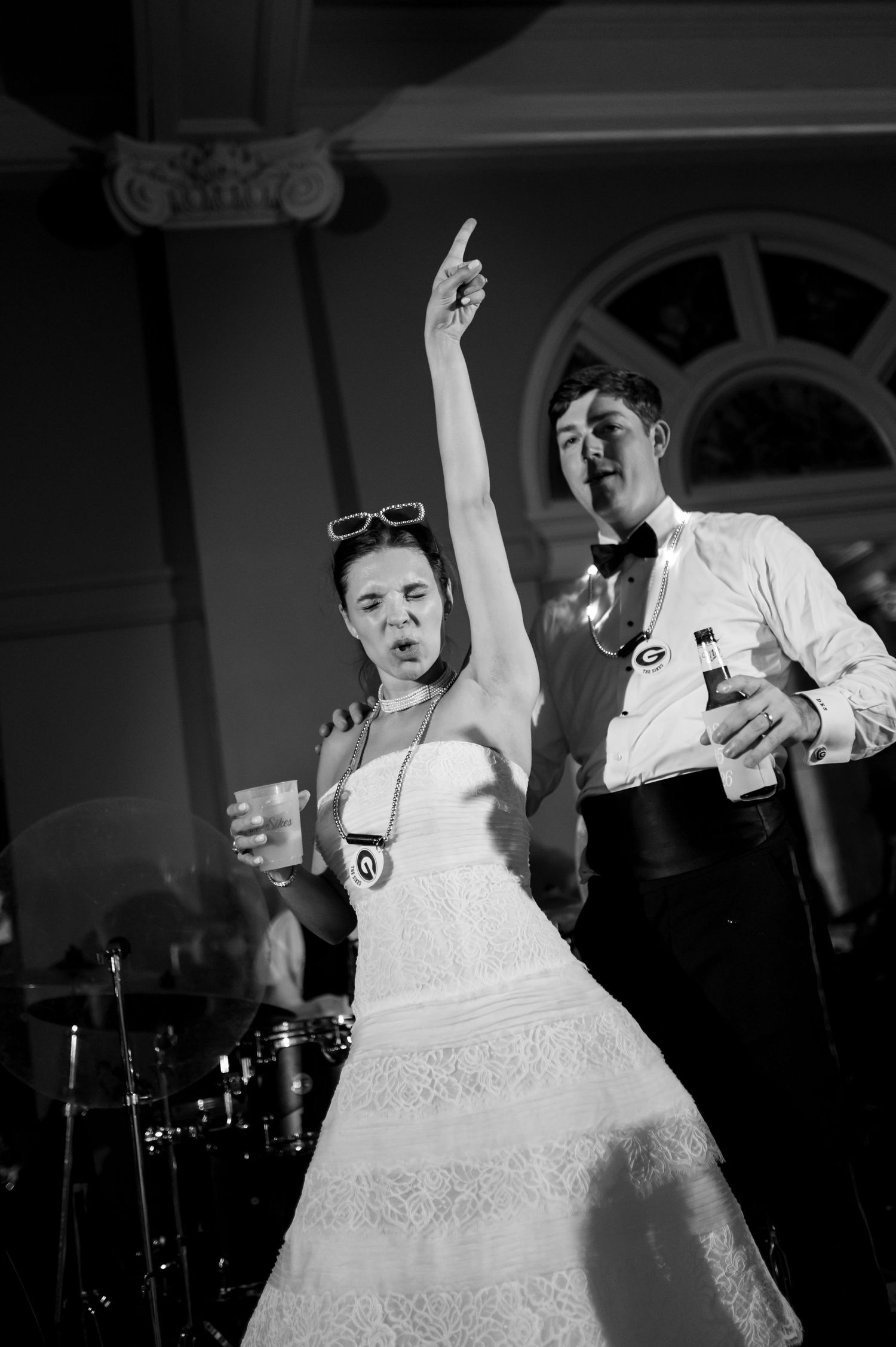 Bride raises arm dancing at wedding reception while groom holds drink in black and white photo.