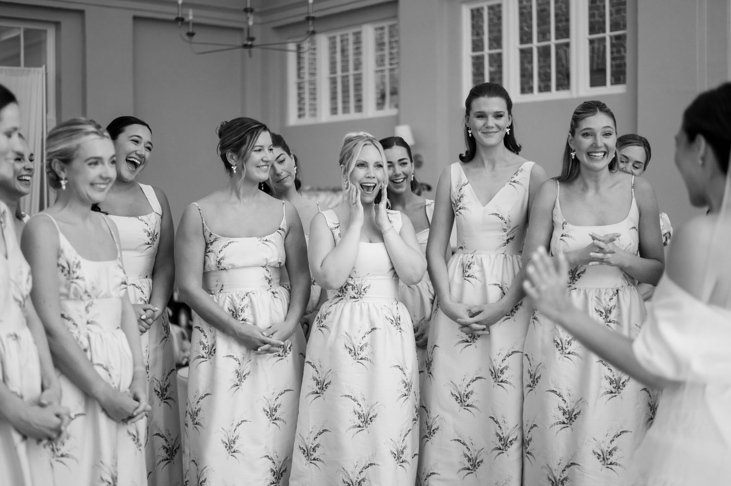 Bridesmaids in matching floral dresses laugh and react emotionally during a wedding preparation moment indoors.
